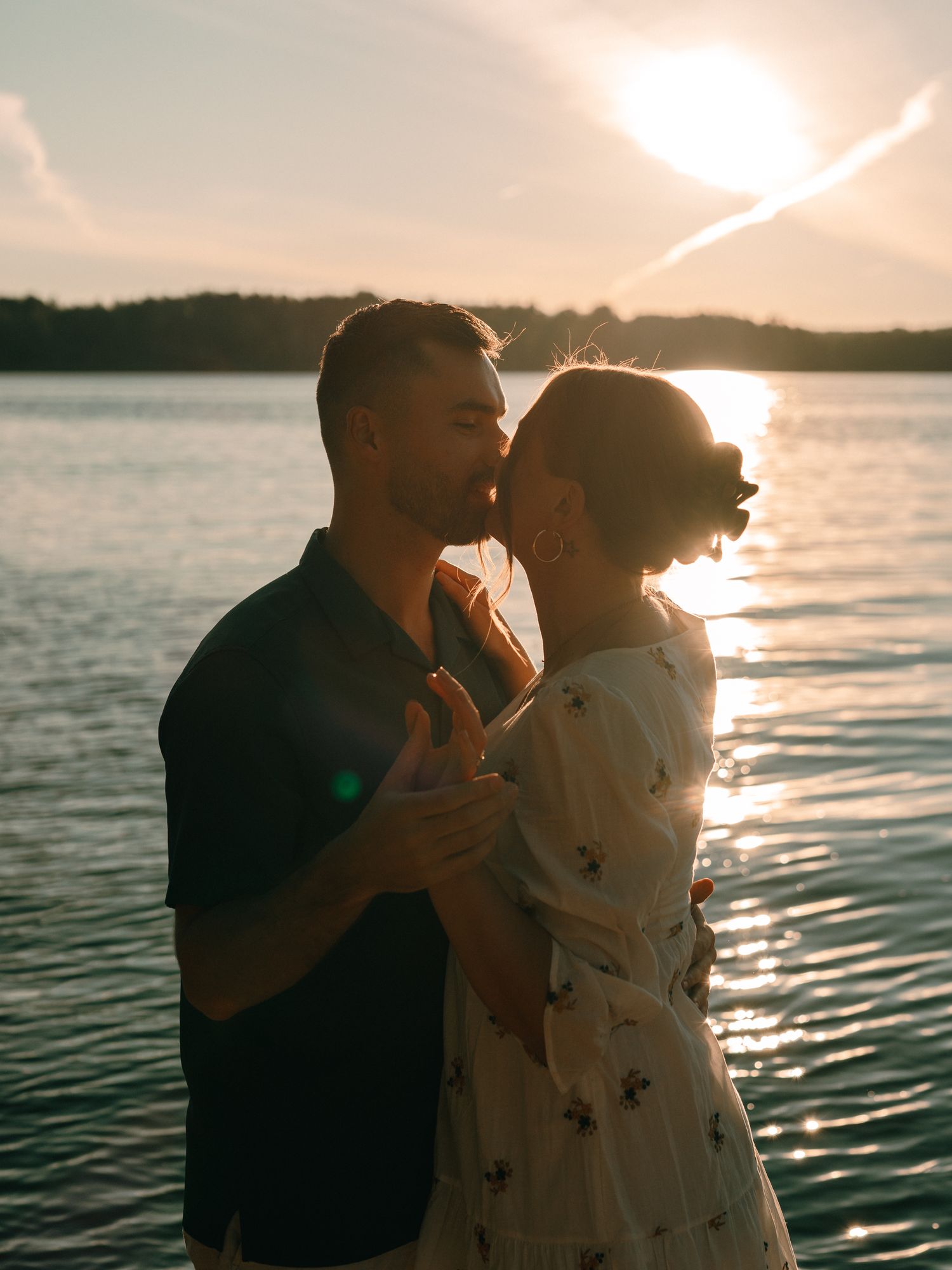 A couple embraces while dancing at sunset by a tranquil lake with golden sunlight streaming through the sky.