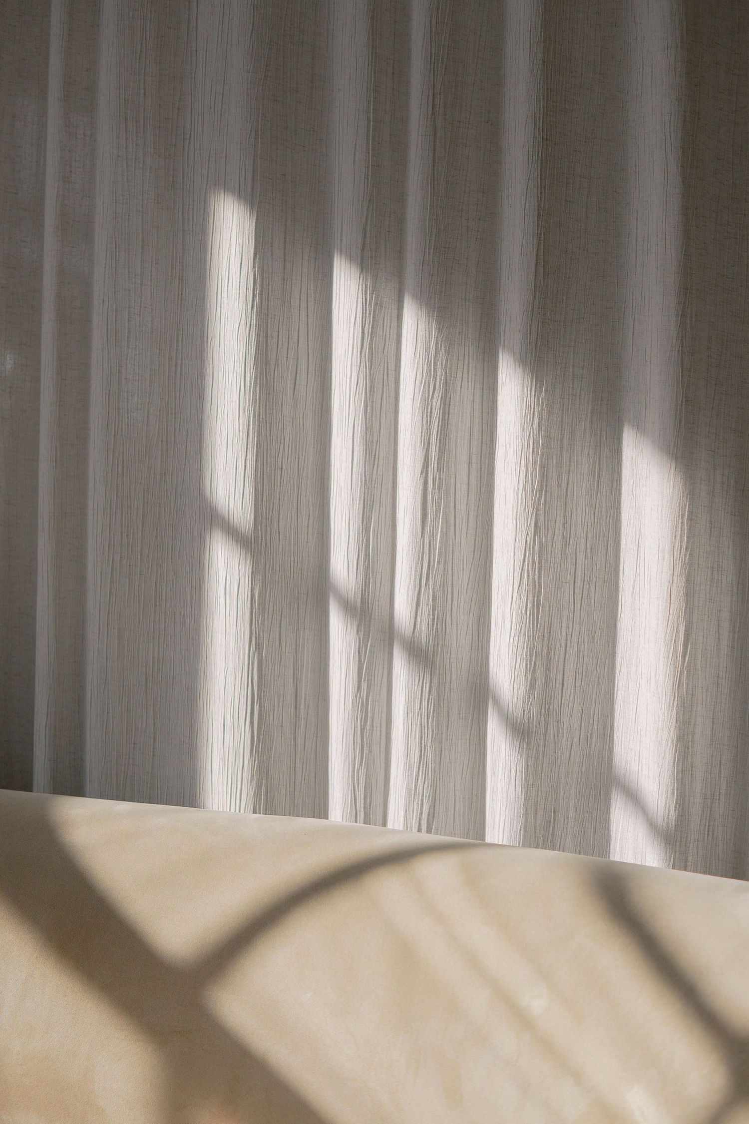 Sunlight casting geometric window shadows on white curtains and beige bedding creating a minimalist abstract pattern.