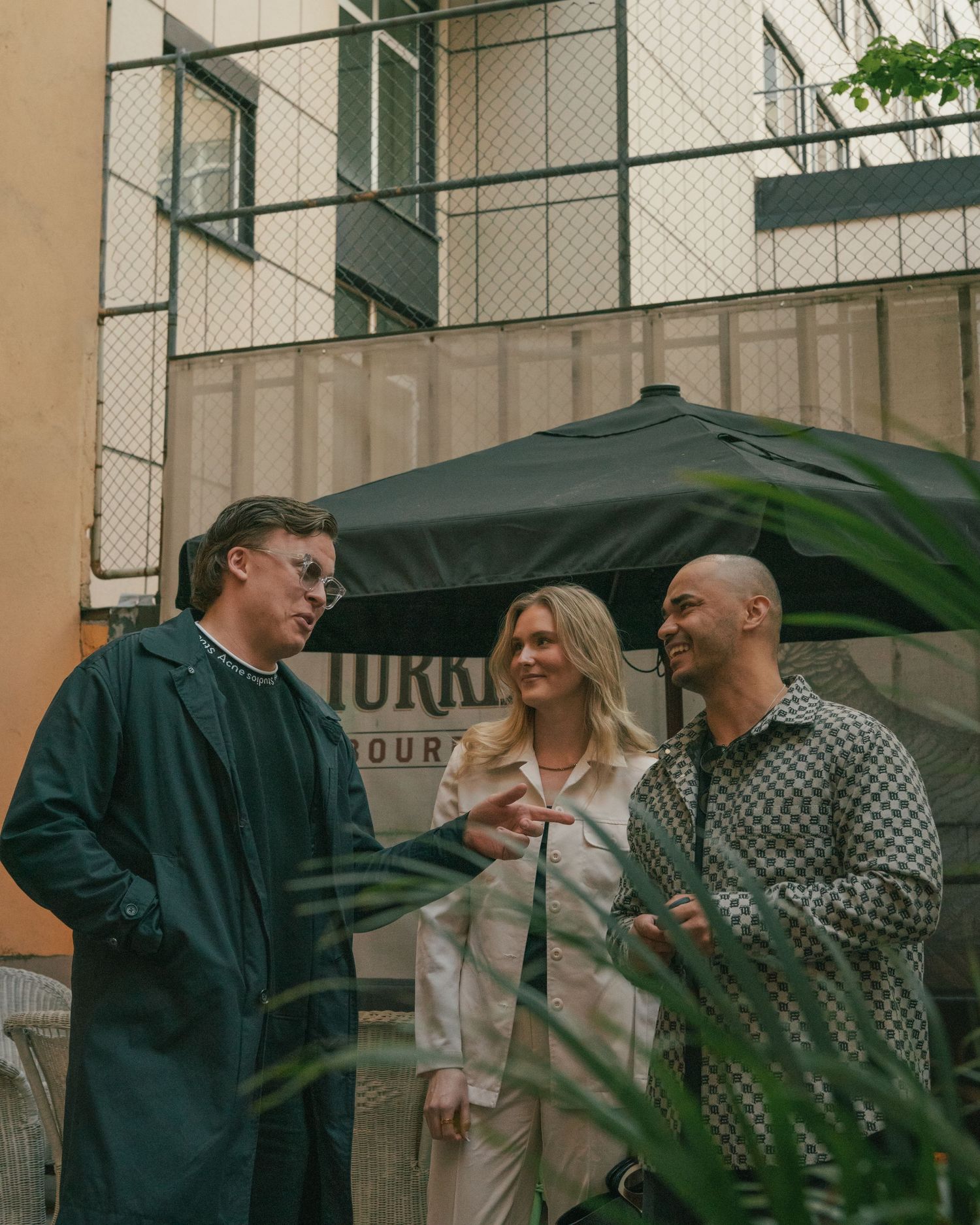 Three people having a casual conversation outdoors near a green umbrella and building wall.