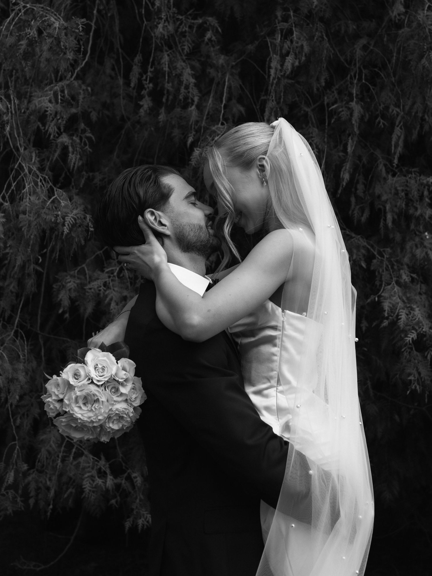 Black and white wedding photo of bride and groom sharing a kiss, bride holding bouquet, veil flowing.