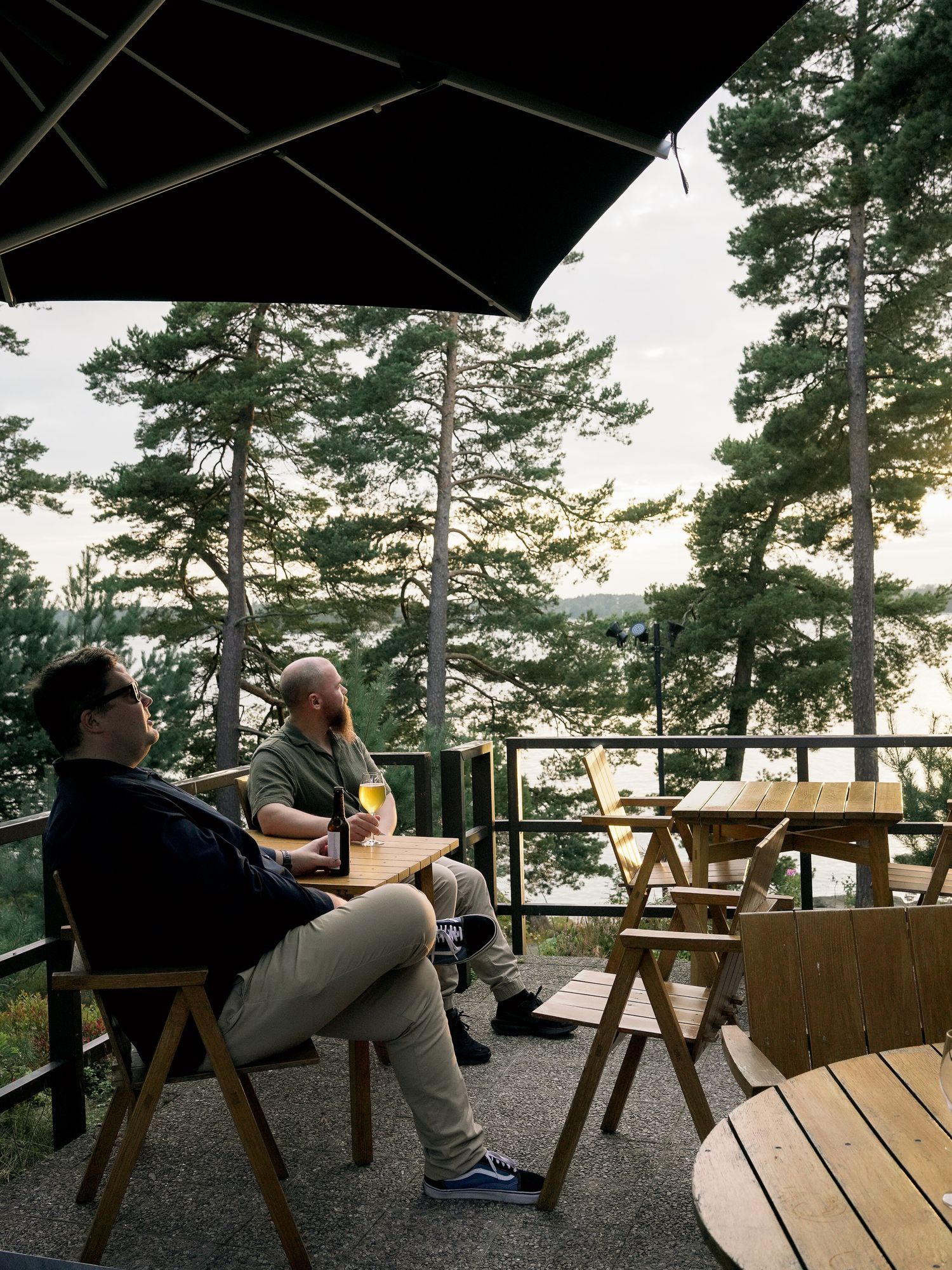 Two men relaxing on a wooden deck with umbrella, surrounded by tall pine trees overlooking a scenic lake view.