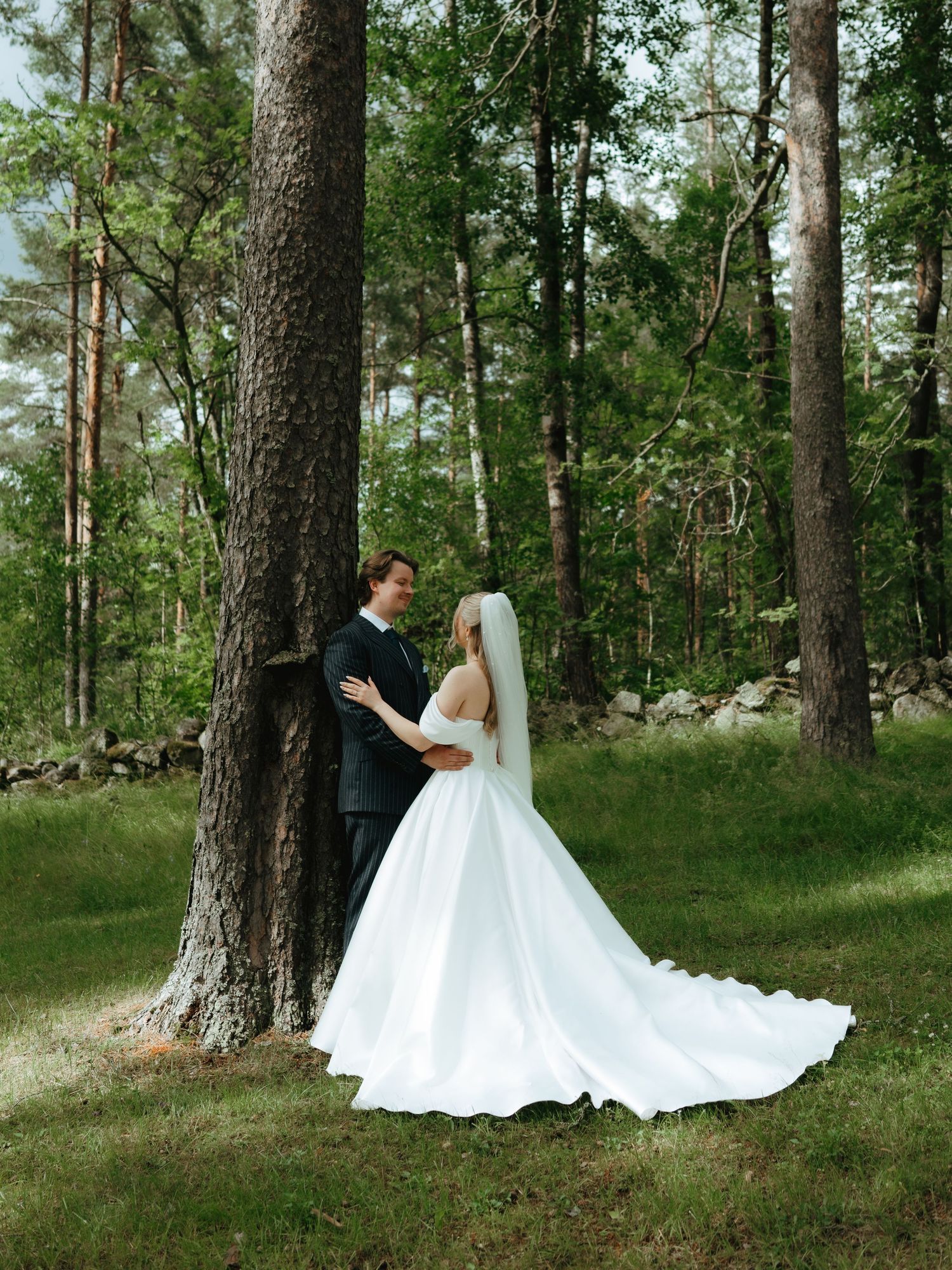 A bride in a white gown and groom in a dark suit embrace by a tree in a lush green forest setting.