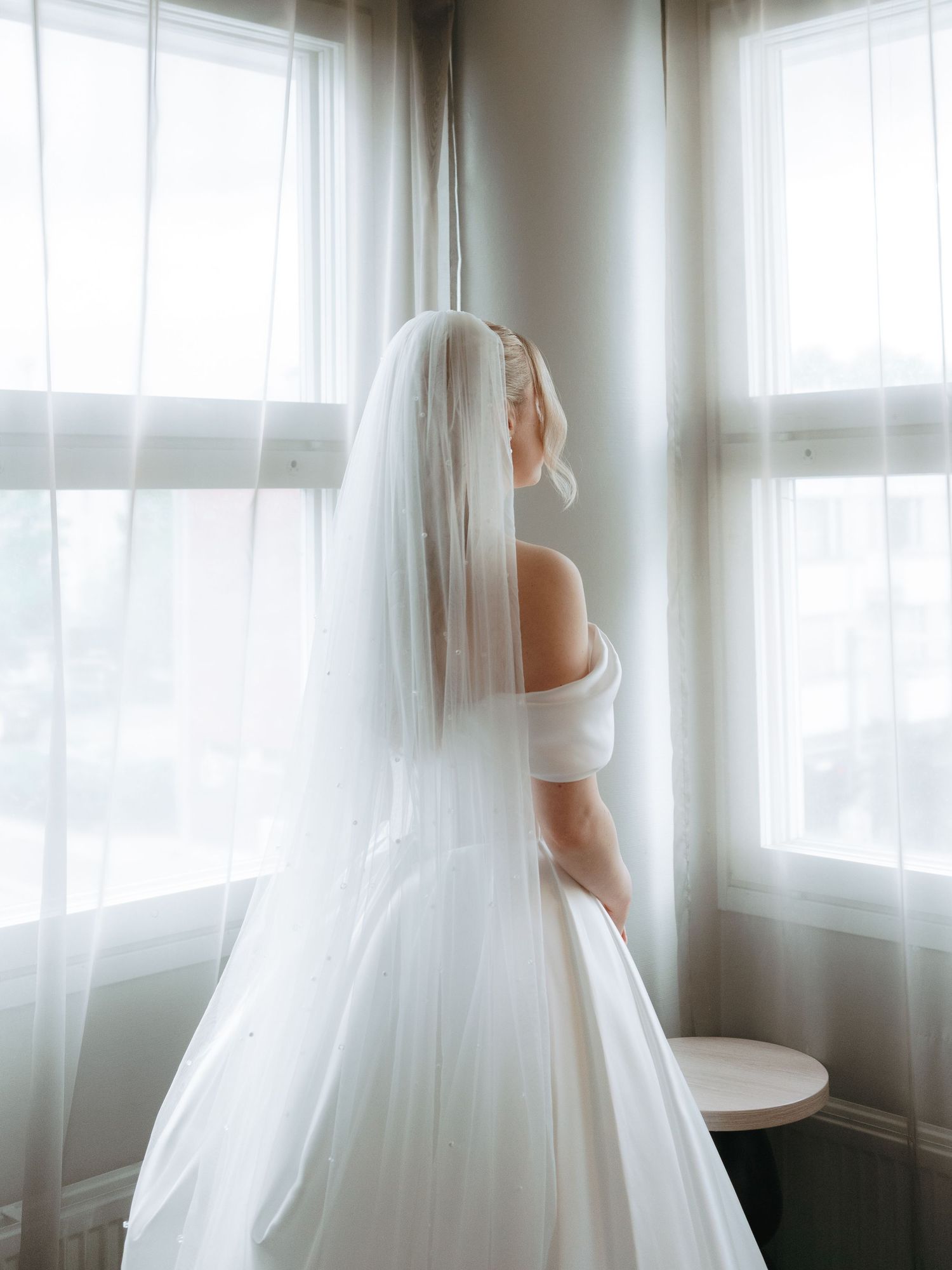 Bride in white off-shoulder gown and long veil stands by sunlit windows, gazing outside on her wedding day.