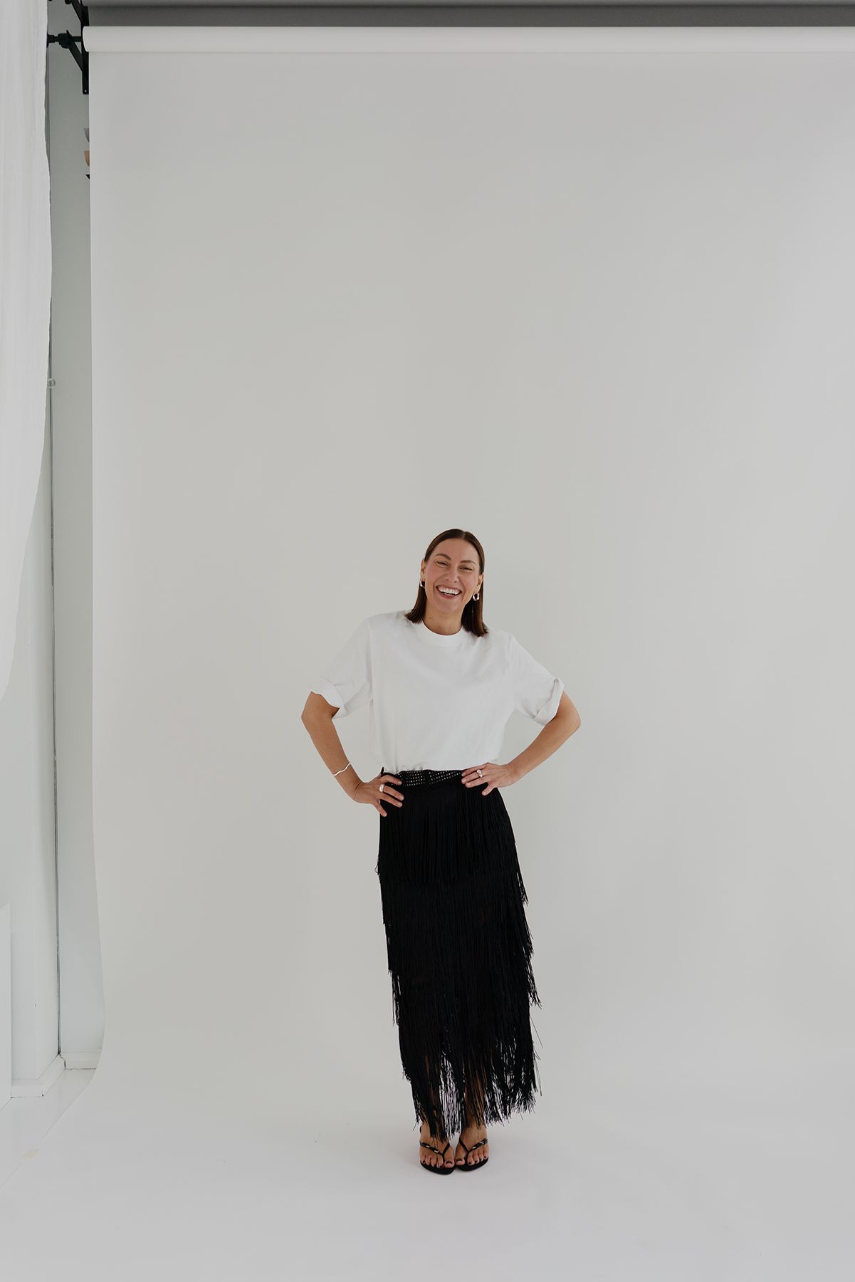 Minimal fashion look with white t-shirt and black fringe skirt against white background.