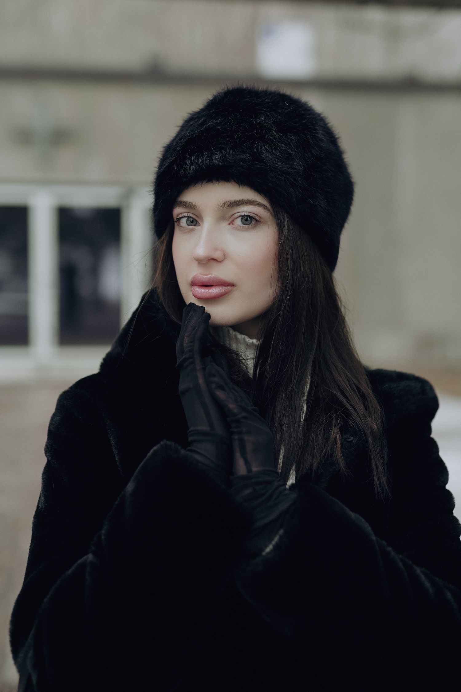 A winter portrait of someone wearing a black fur hat, gloves and coat against a soft blurred background.