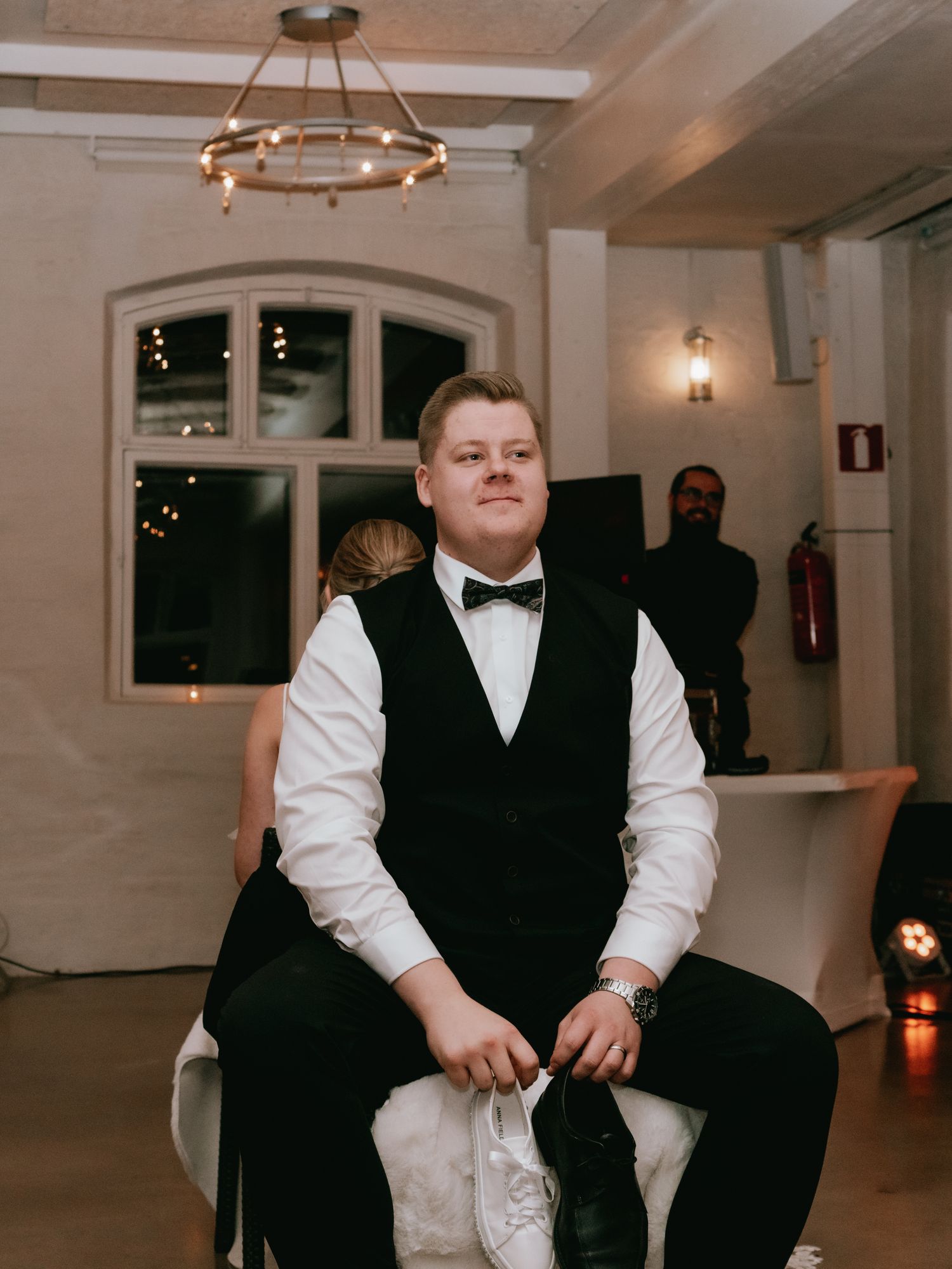 A person in formal attire with black vest and bowtie sits on a light-colored chair in an elegant venue with chandeliers.