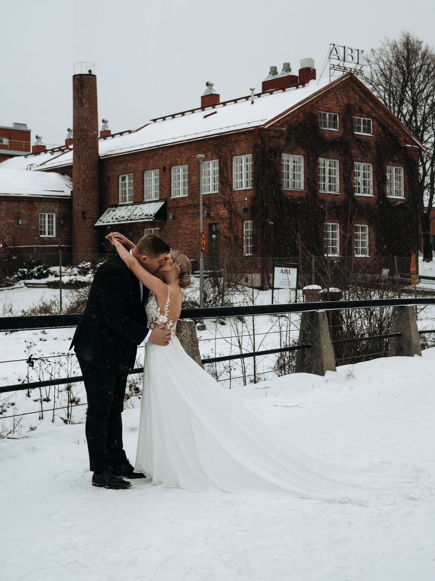 A couple in wedding attire share a romantic moment in the snow outside a historic brick building in winter.