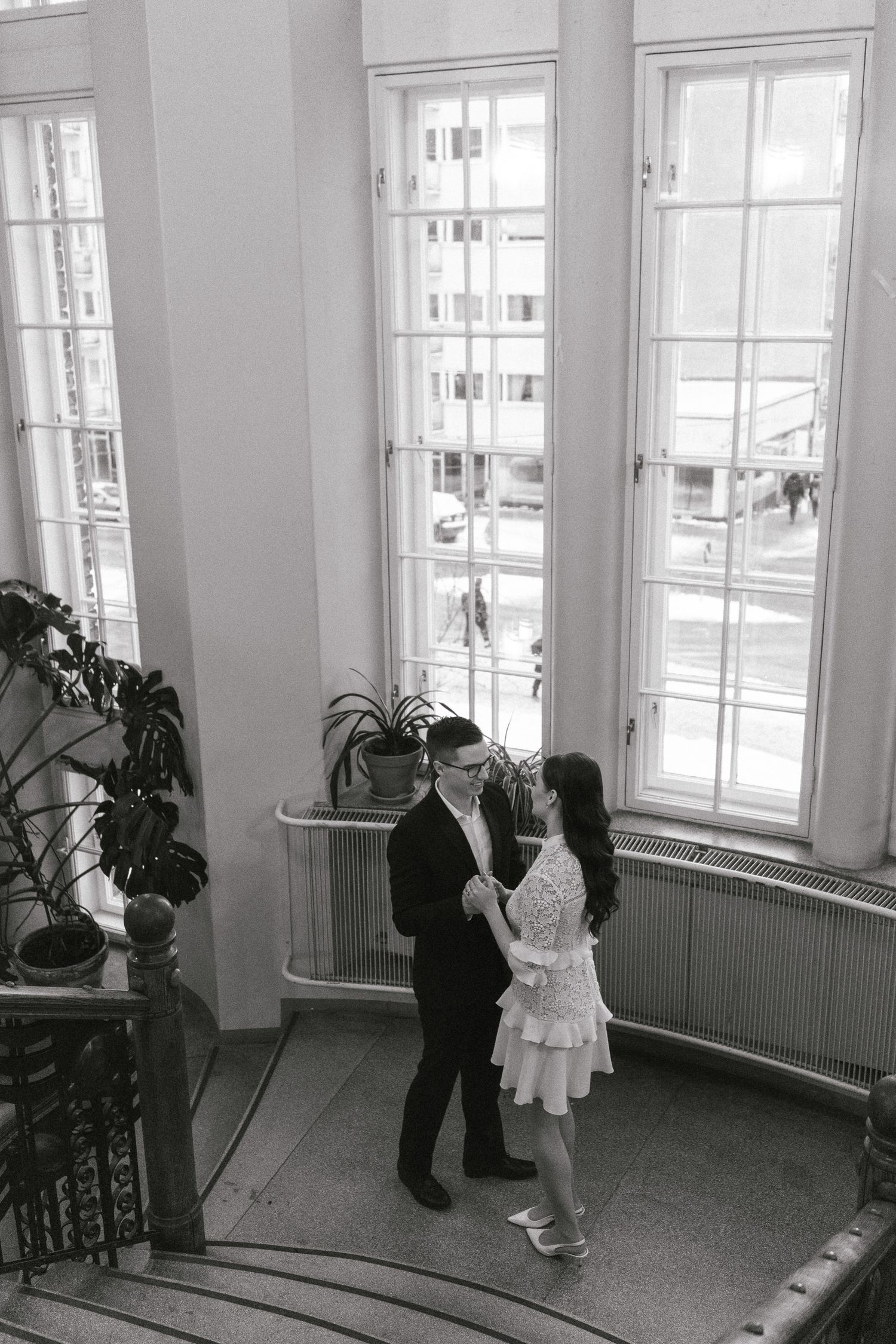Two figures stand in an intimate moment by tall vintage windows in a black and white photograph.