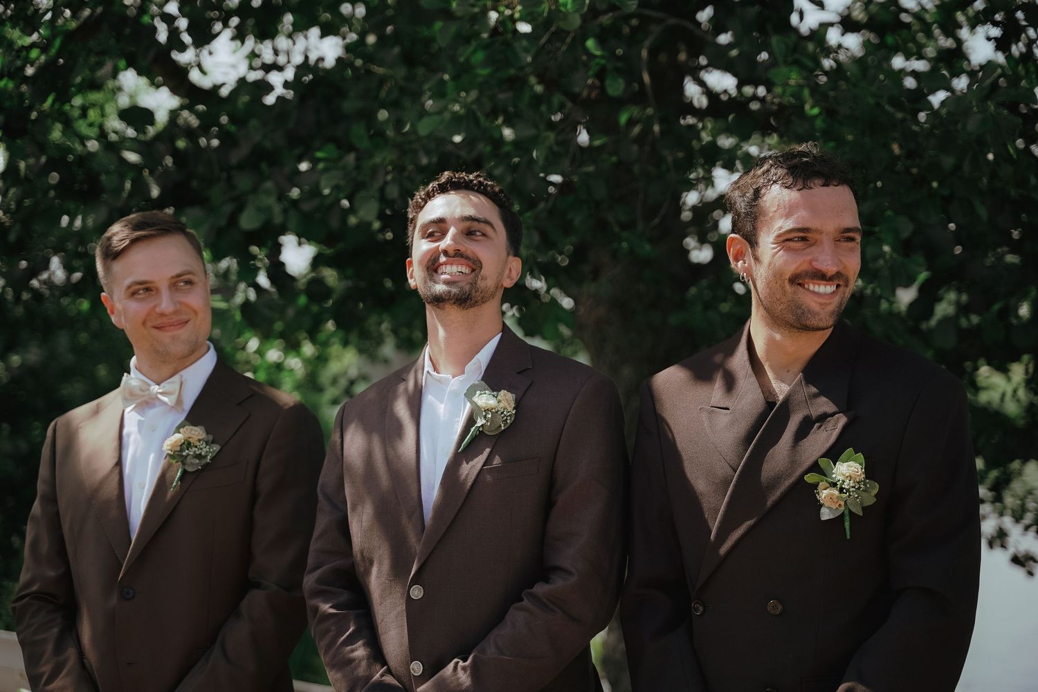 Three groomsmen in dark suits with boutonnieres stand together outdoors with greenery in background.