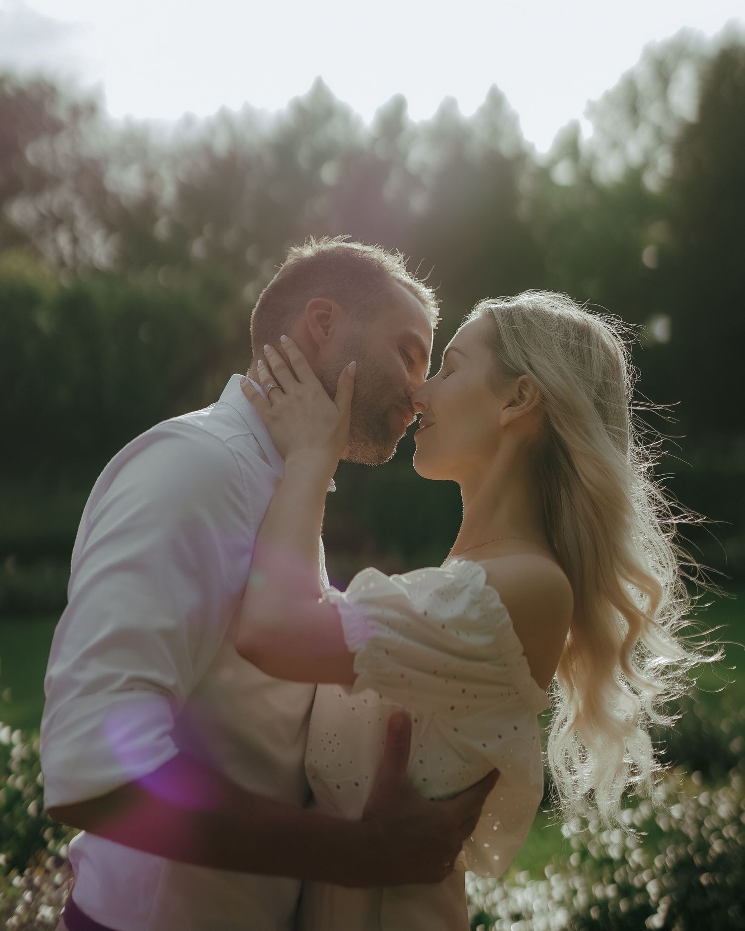 A couple shares a romantic embrace and kiss in golden sunset light among greenery and trees in nature.