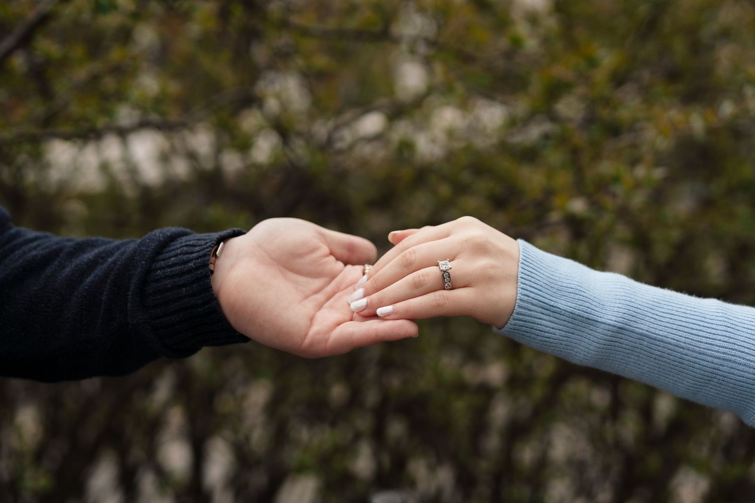 Two hands gently touching, with a diamond engagement ring on the woman's finger, against a blurred green background.