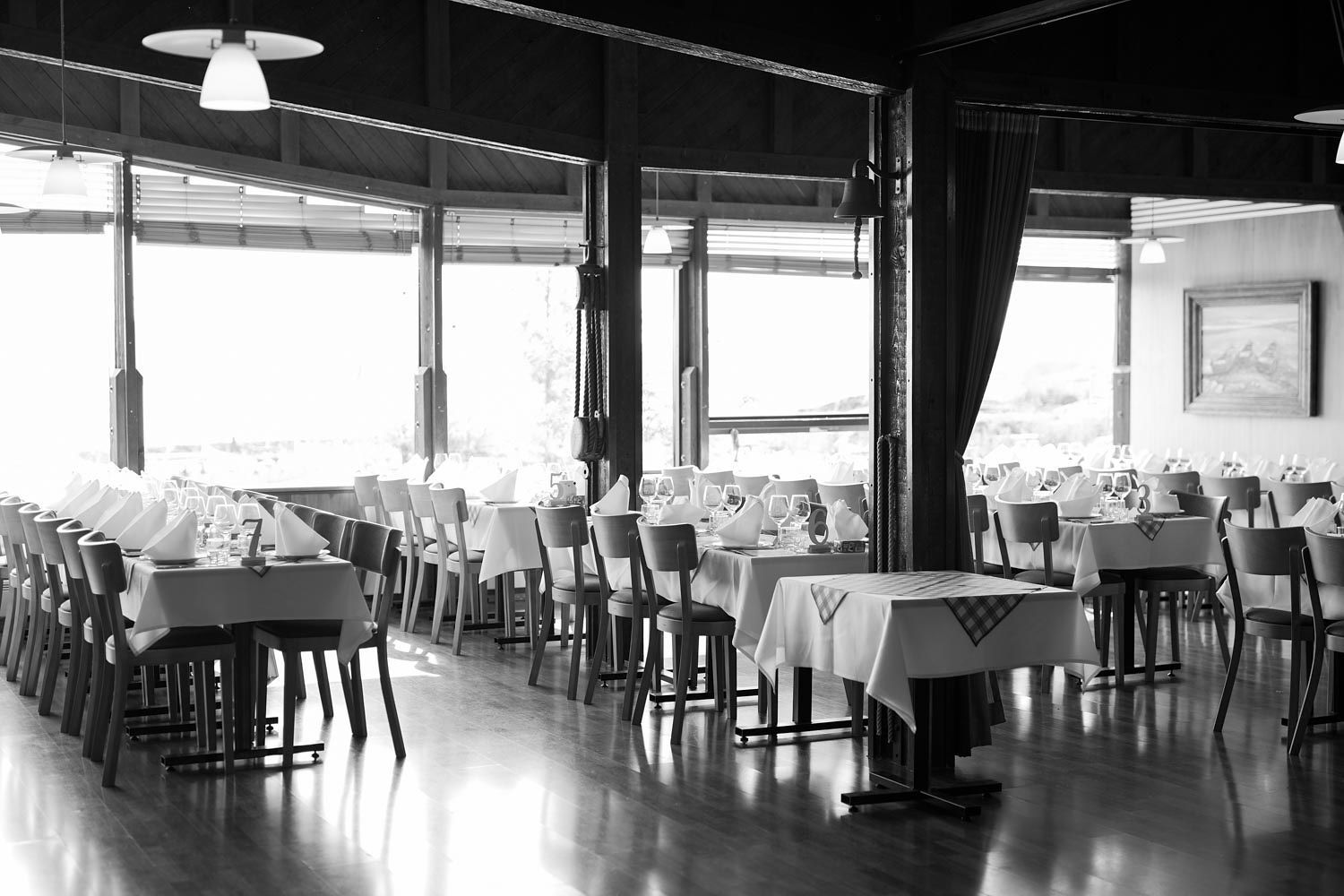 Black and white photo of an elegant empty restaurant dining room with white tablecloths and large windows.