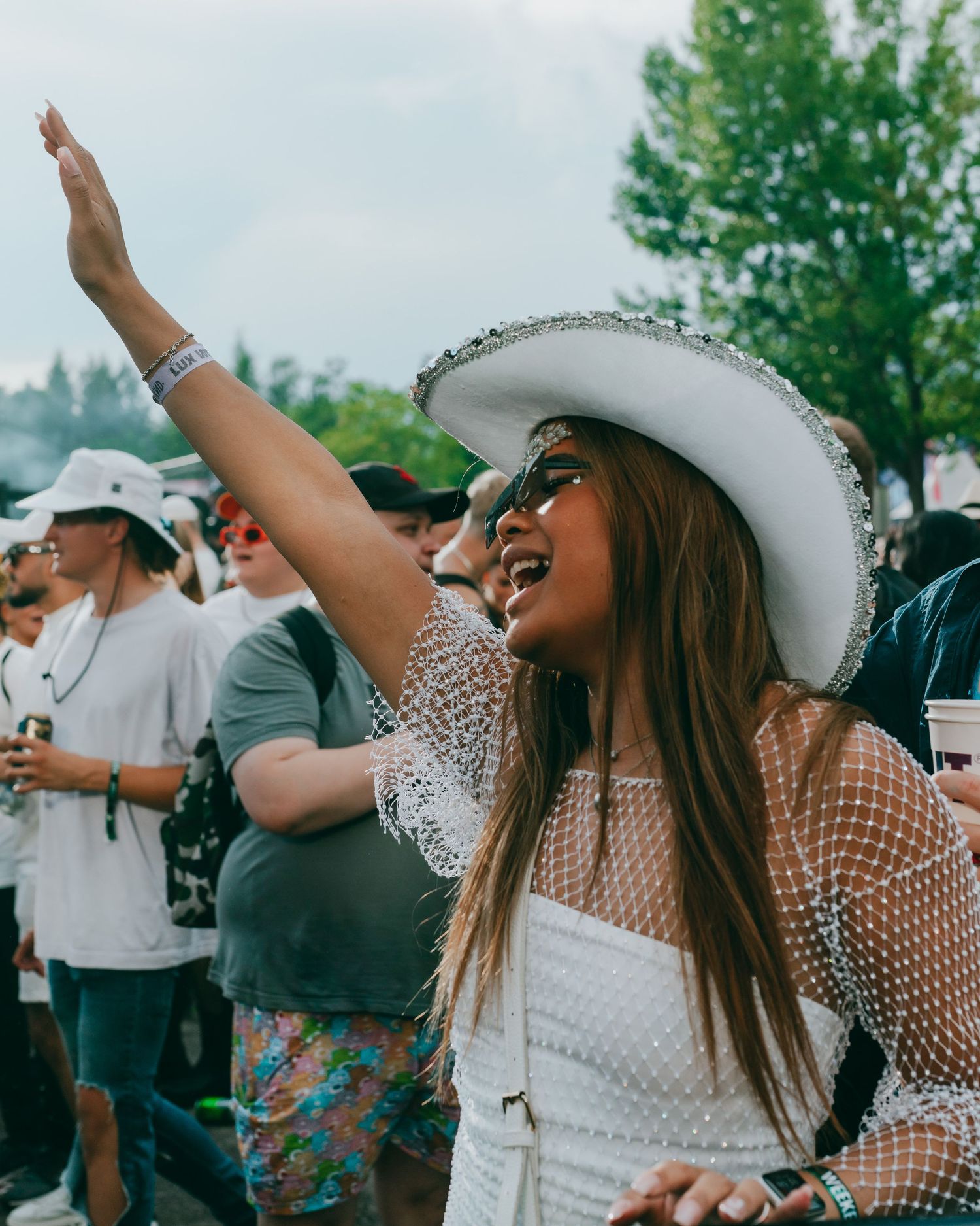 Festival attendee in white cowboy hat and mesh top raises hand joyfully in outdoor crowd.