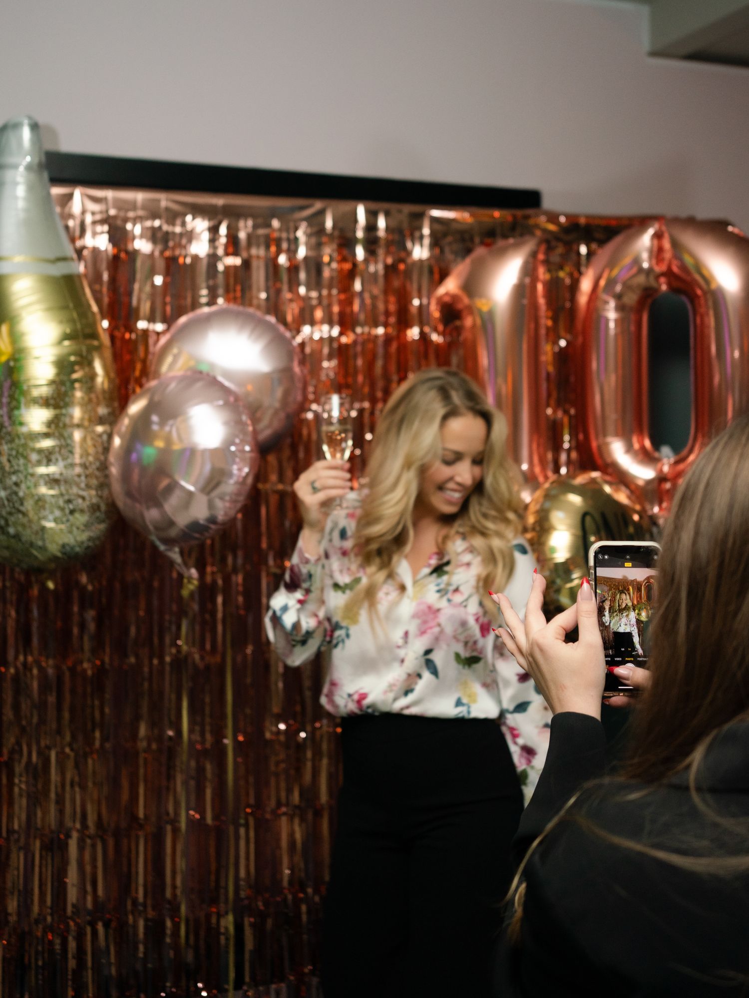 People celebrating at a party with rose gold balloon decorations and champagne against a metallic fringe backdrop.