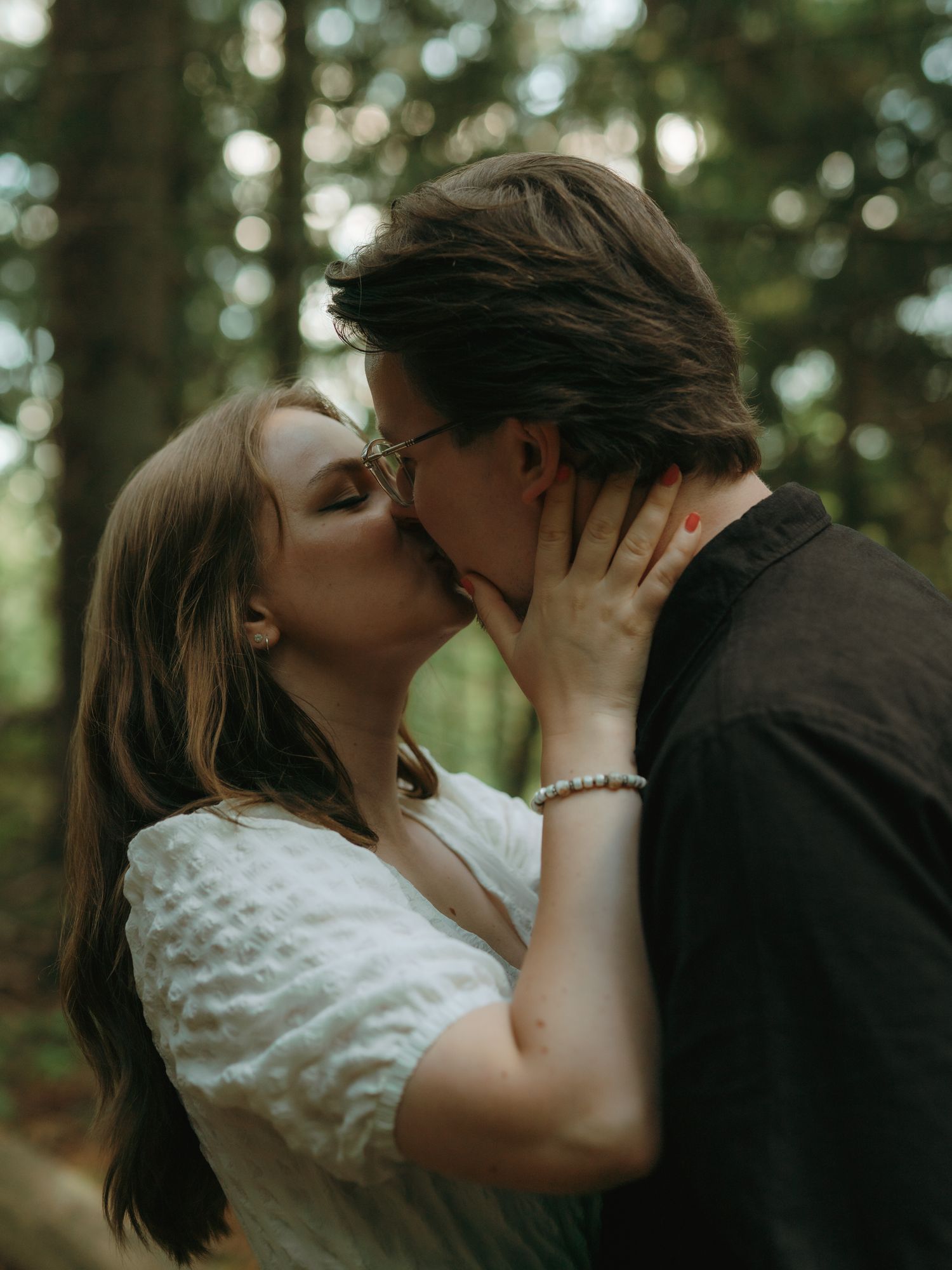 A romantic couple shares an intimate kiss in a dreamy forest setting with soft bokeh lighting in the background.