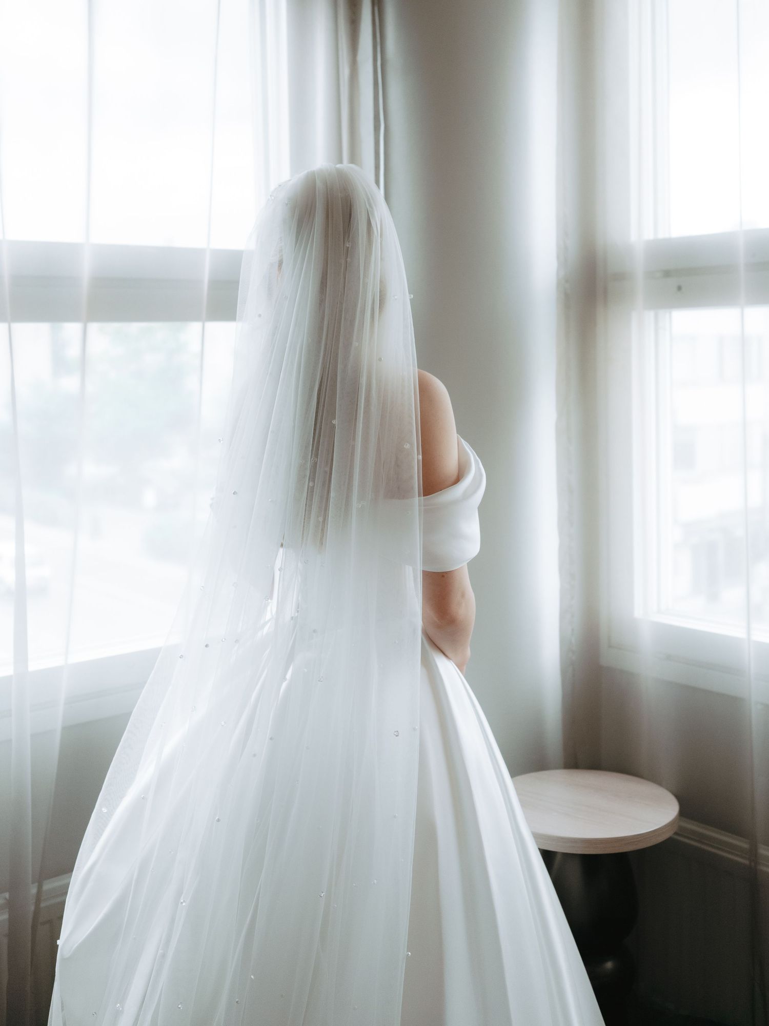 Bride in white wedding gown and veil stands facing window in soft natural light.
