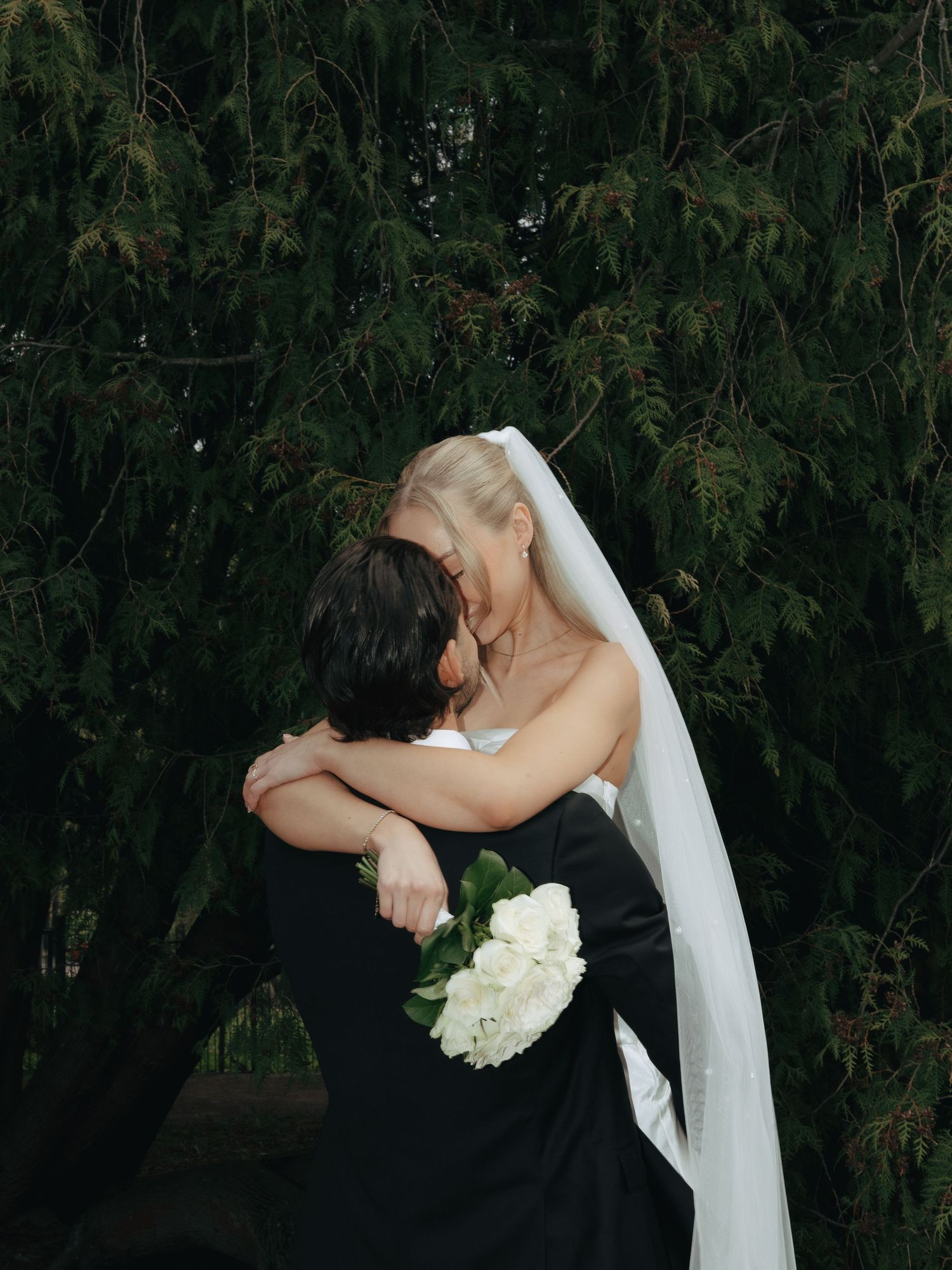 Bride in white veil holding bouquet embraces groom in black suit outdoors near lush green trees.