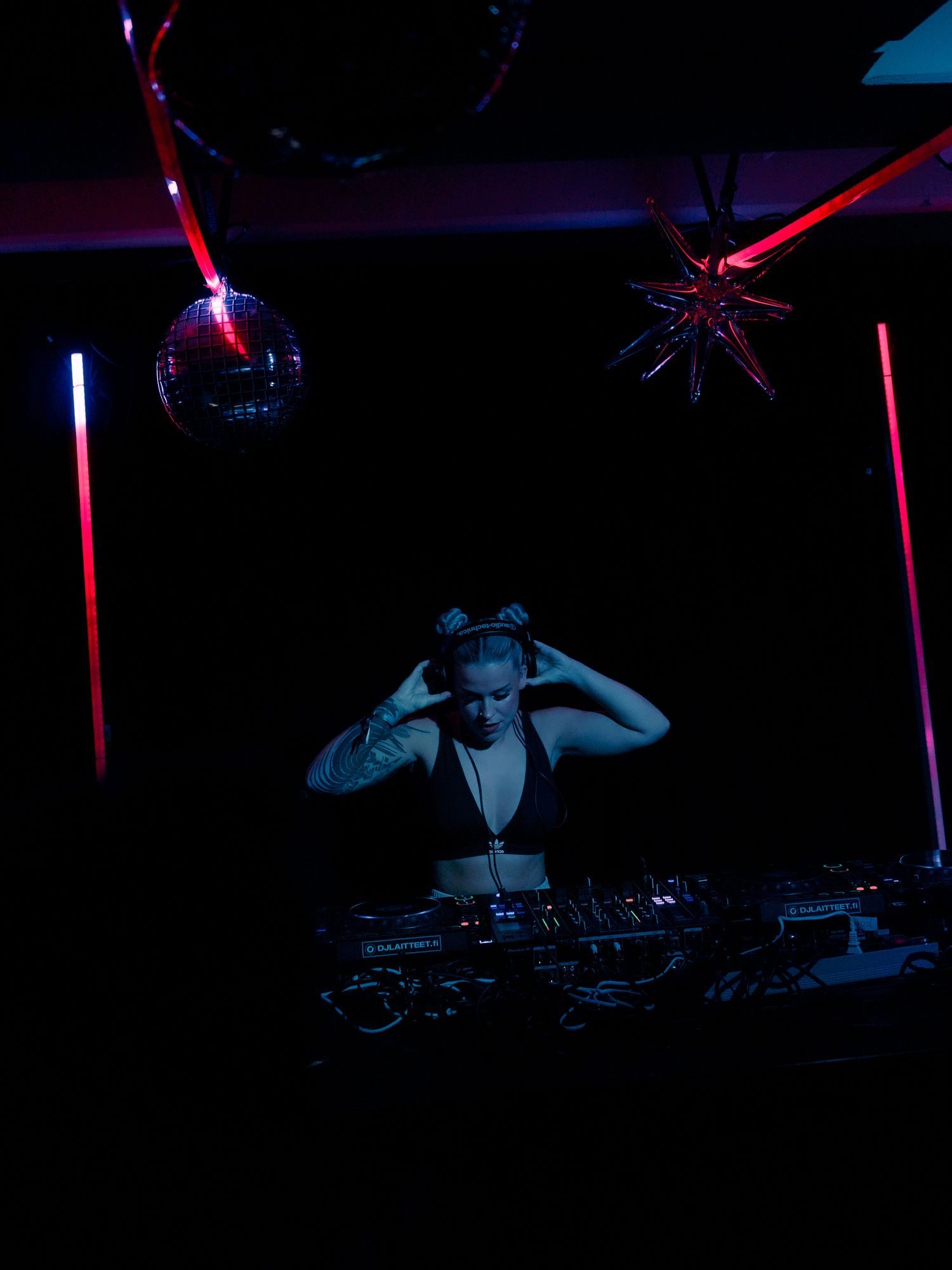 A DJ performs at a nightclub under moody blue and red lighting with disco balls and equipment visible.