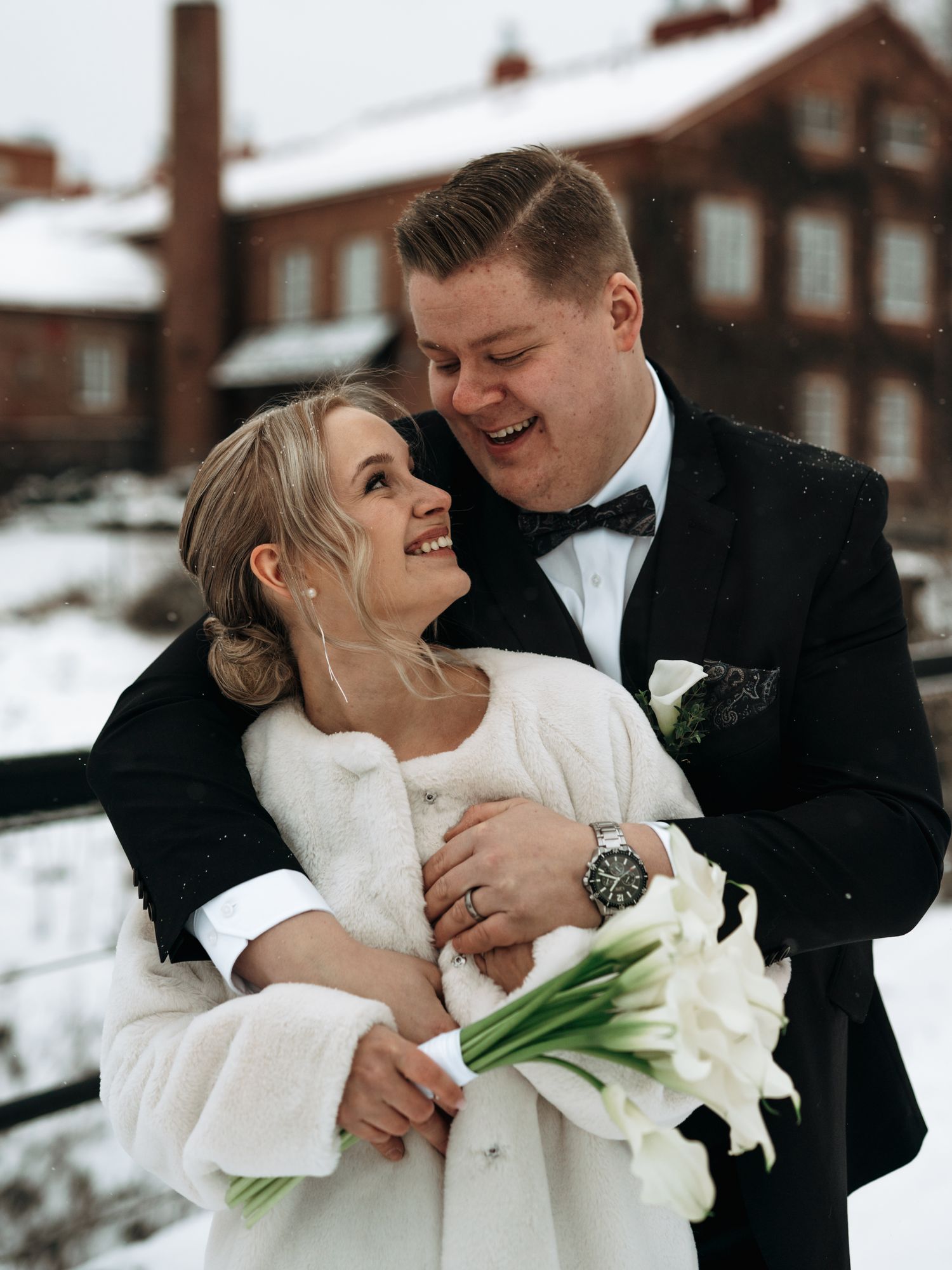 A joyful winter wedding couple embraces while wearing formal attire and holding white calla lilies in the snow.