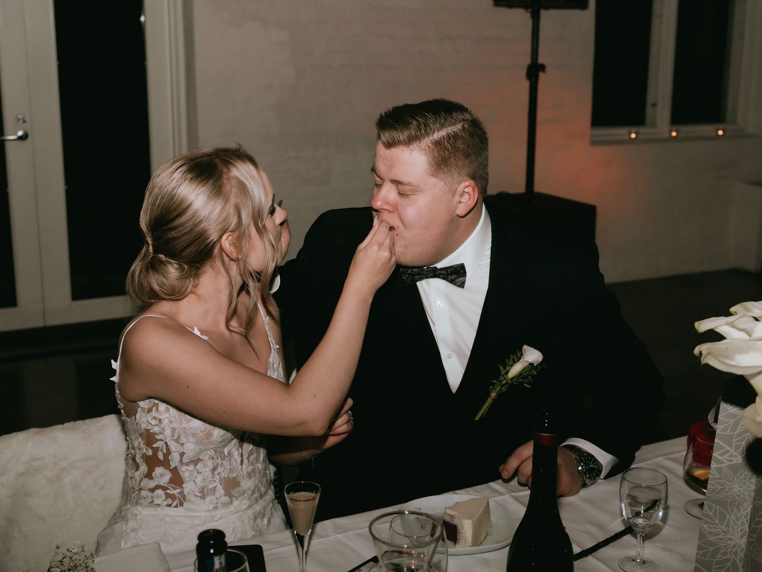 A couple shares a playful moment at their wedding reception, seated at a table with a white tablecloth and calla lily decor.