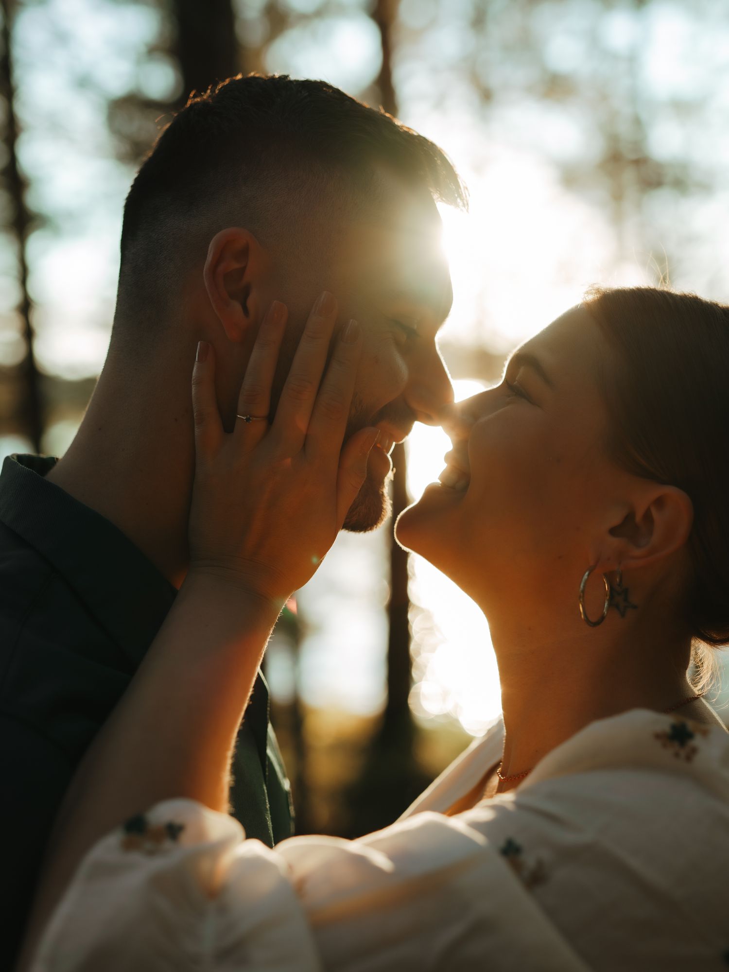 Silhouetted couple sharing an intimate moment with sun flare through trees in romantic backlit portrait.