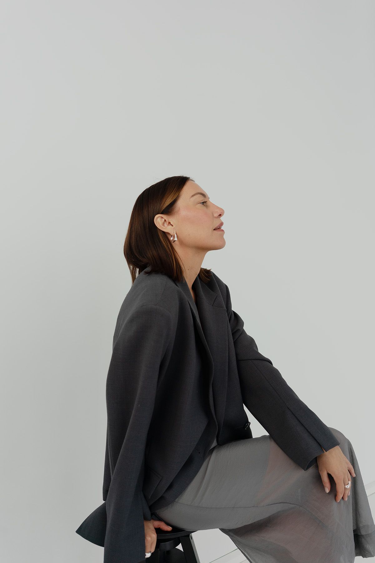 Model wearing dark gray blazer sits in contemplative pose against light background in minimalist fashion shoot.