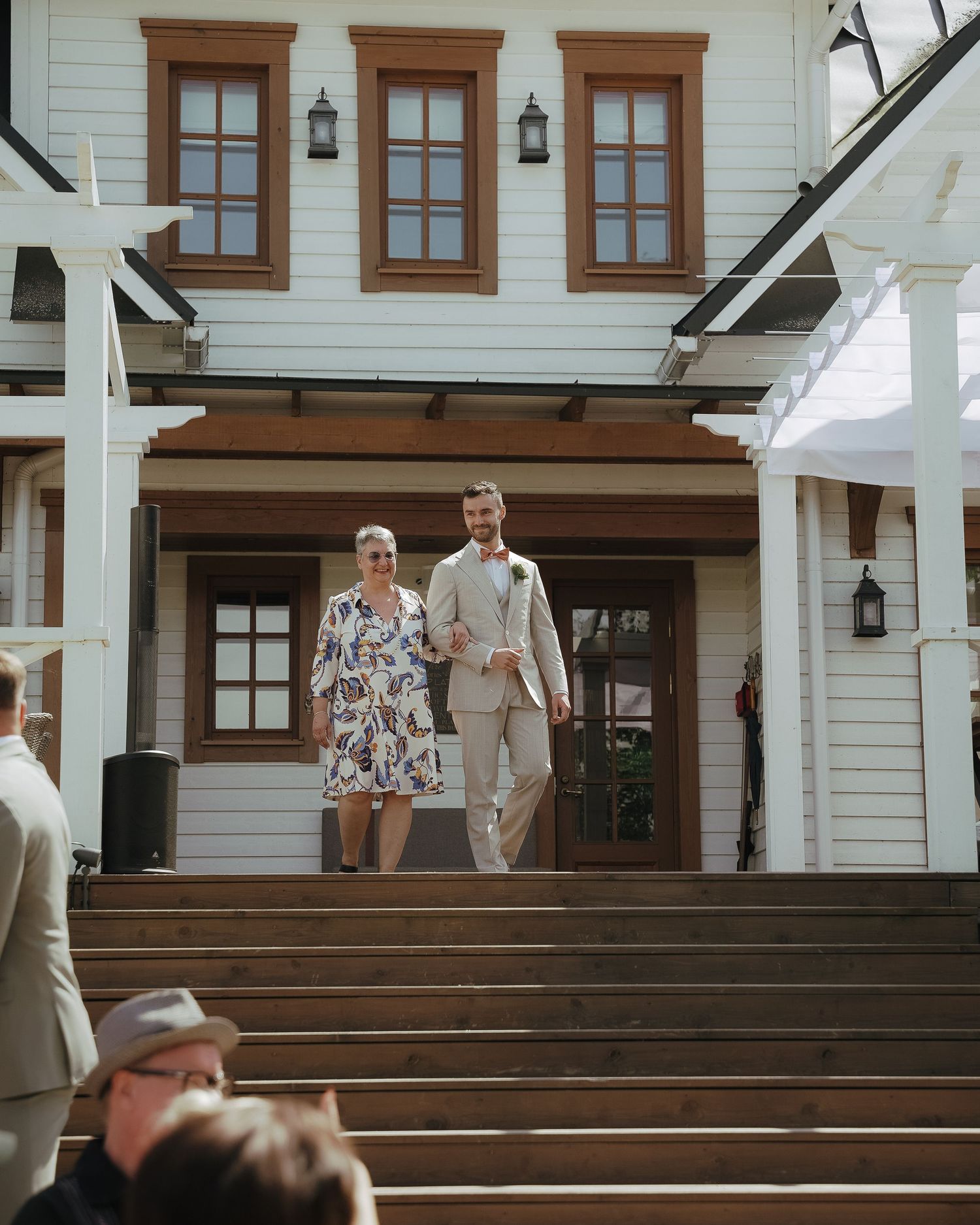 A couple descends wooden stairs at a beautiful white farmhouse with brown trim windows and black lanterns.
