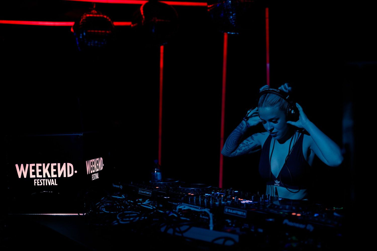 DJ performs at Weekend Festival under blue and red stage lighting with equipment setup visible in dark nightclub setting.