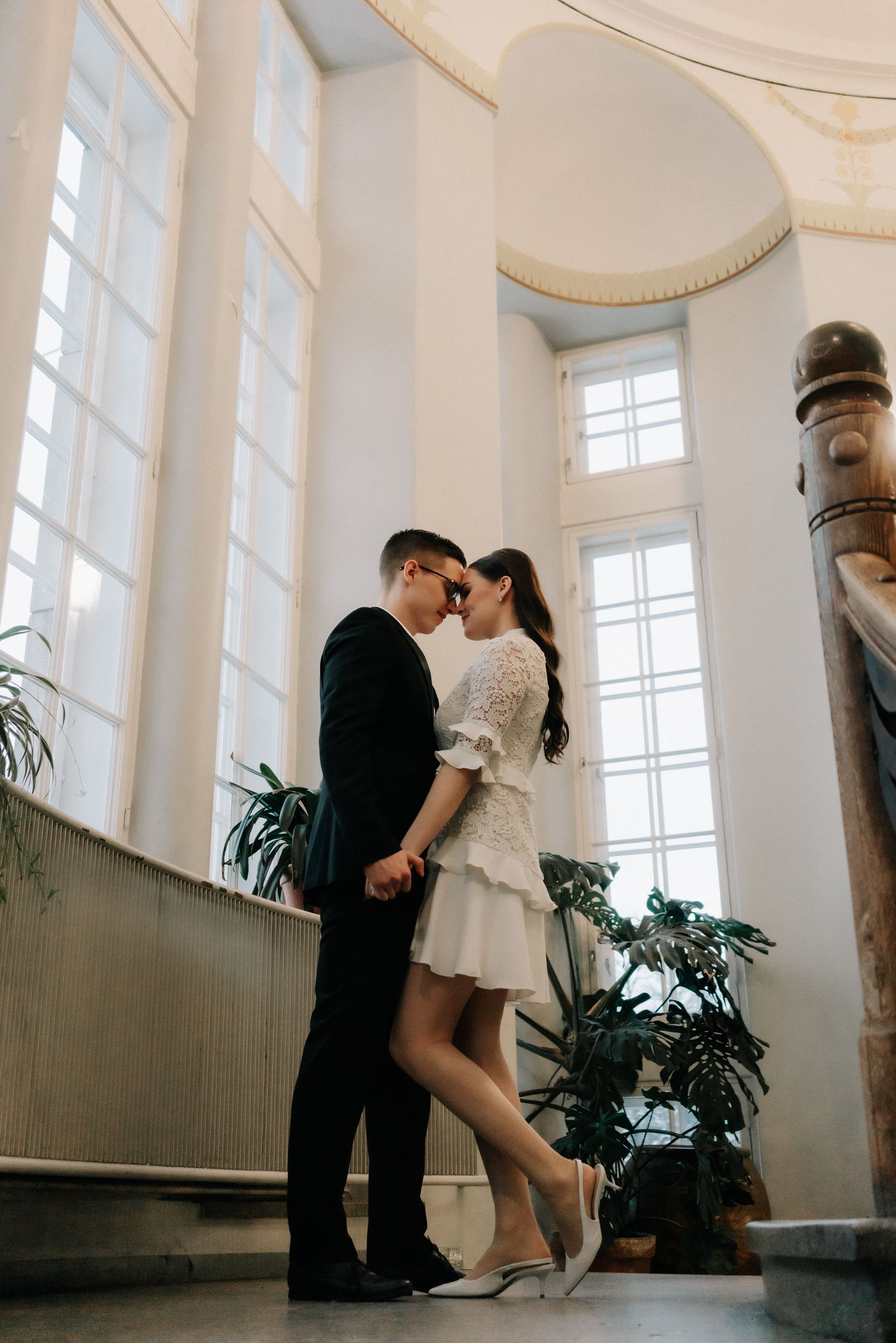 Romantic couple in wedding attire share an intimate moment in an elegant interior with tall arched windows and potted plants.