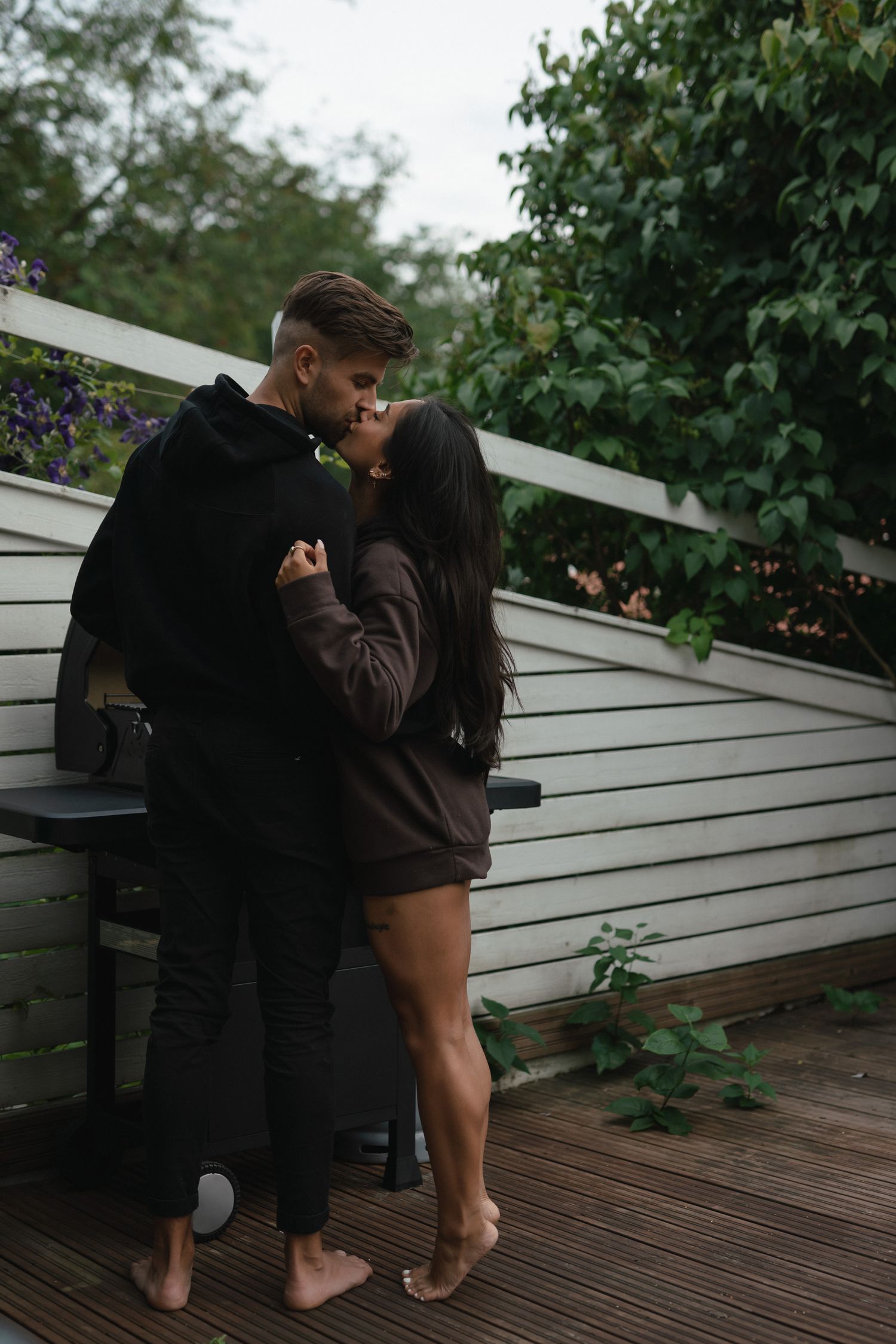 A couple shares an intimate moment on a wooden deck against white siding, surrounded by green foliage.