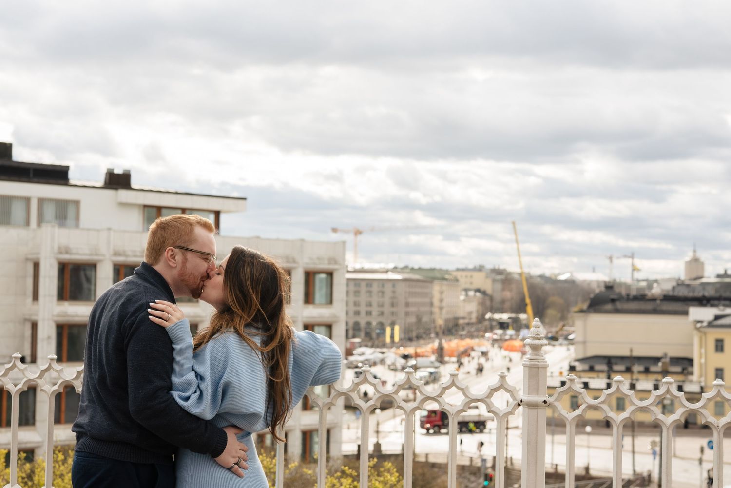 Couple sharing a romantic kiss on a rooftop terrace with a cloudy city skyline backdrop.