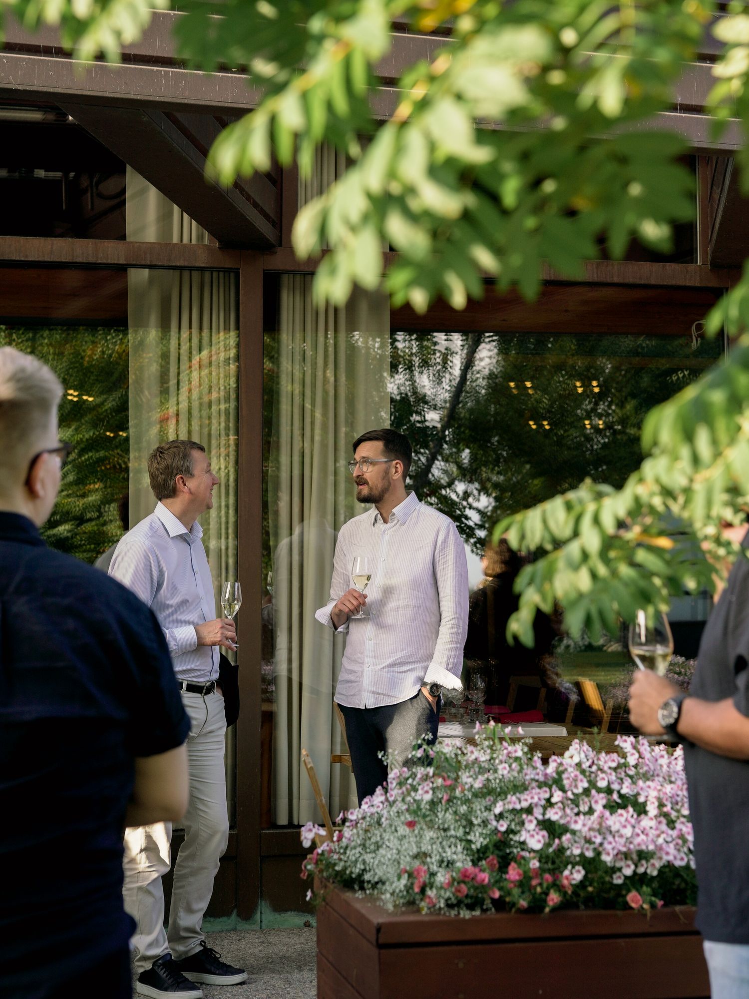 Men in casual attire socializing outdoors at an elegant garden event, holding champagne glasses near blooming flowers.