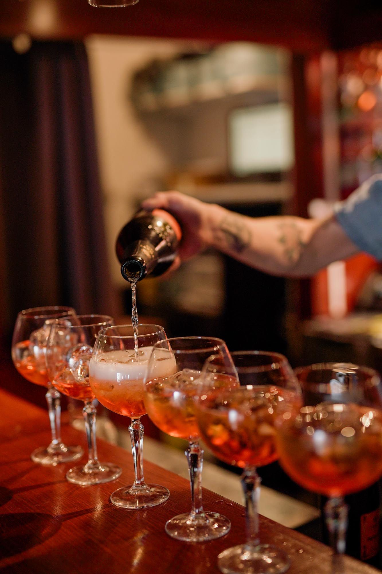 Tattooed bartender pouring cocktails into a row of stemmed glasses filled with ice on a wooden bar.