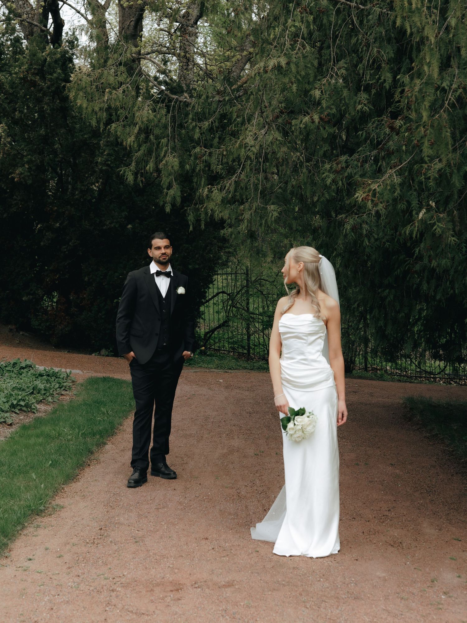 Bride in white strapless gown holding bouquet faces groom in black tuxedo on garden path surrounded by tall evergreen trees.