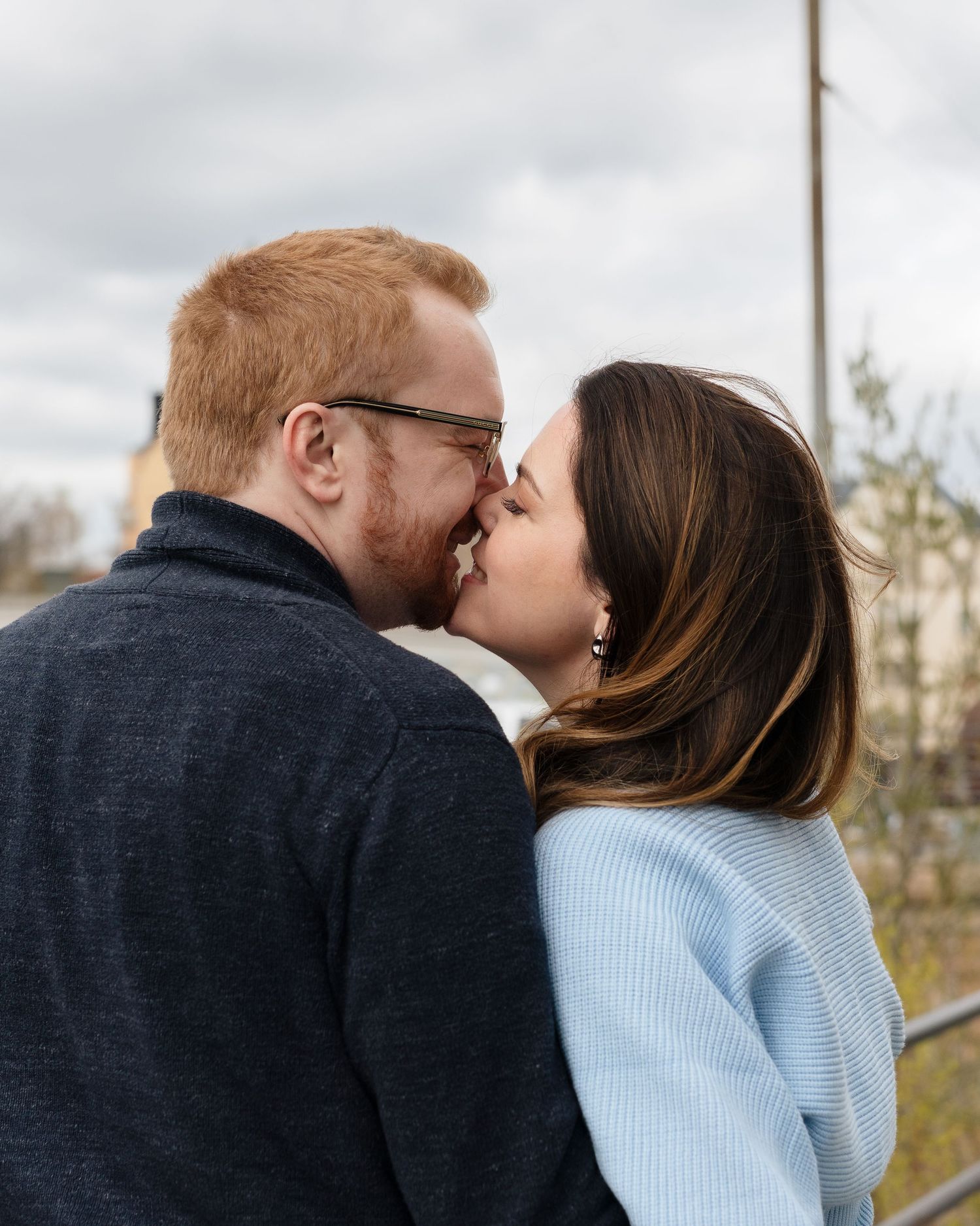 A couple sharing a romantic kiss outdoors, man with red hair and glasses, woman in light blue sweater.