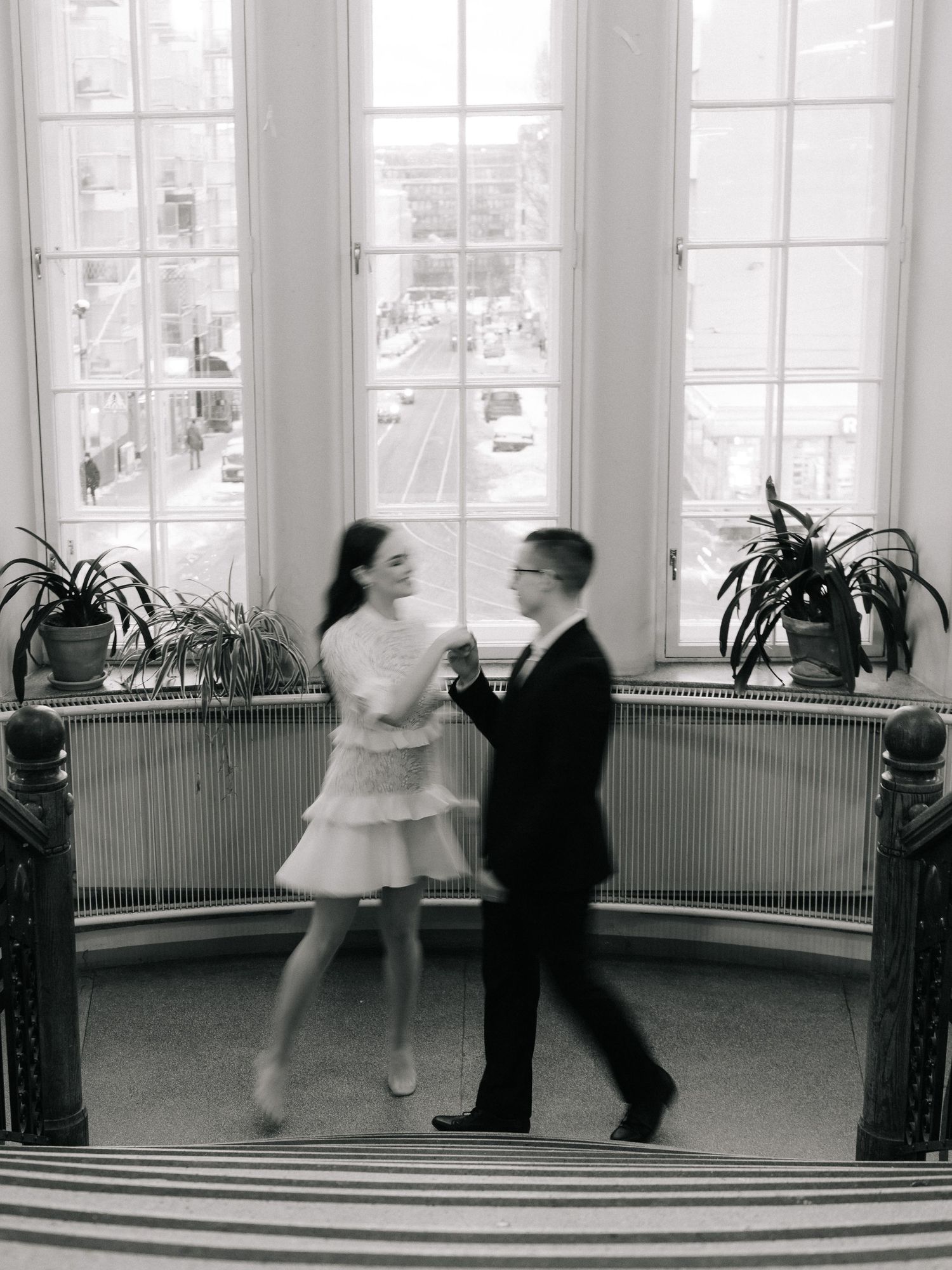 A couple dances together in an elegant bay window area with houseplants, captured in classic black and white photography.