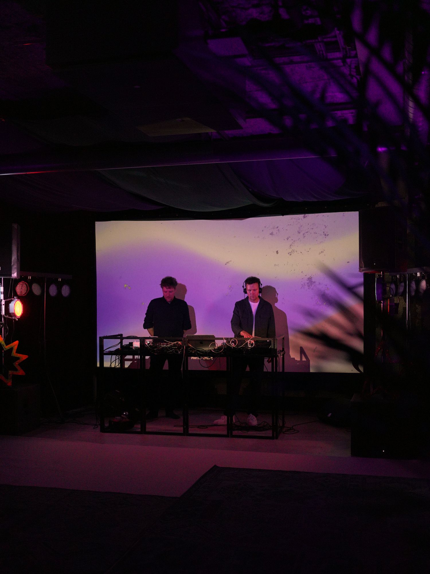 Two DJs performing on stage with purple backdrop lighting at an indoor event venue.