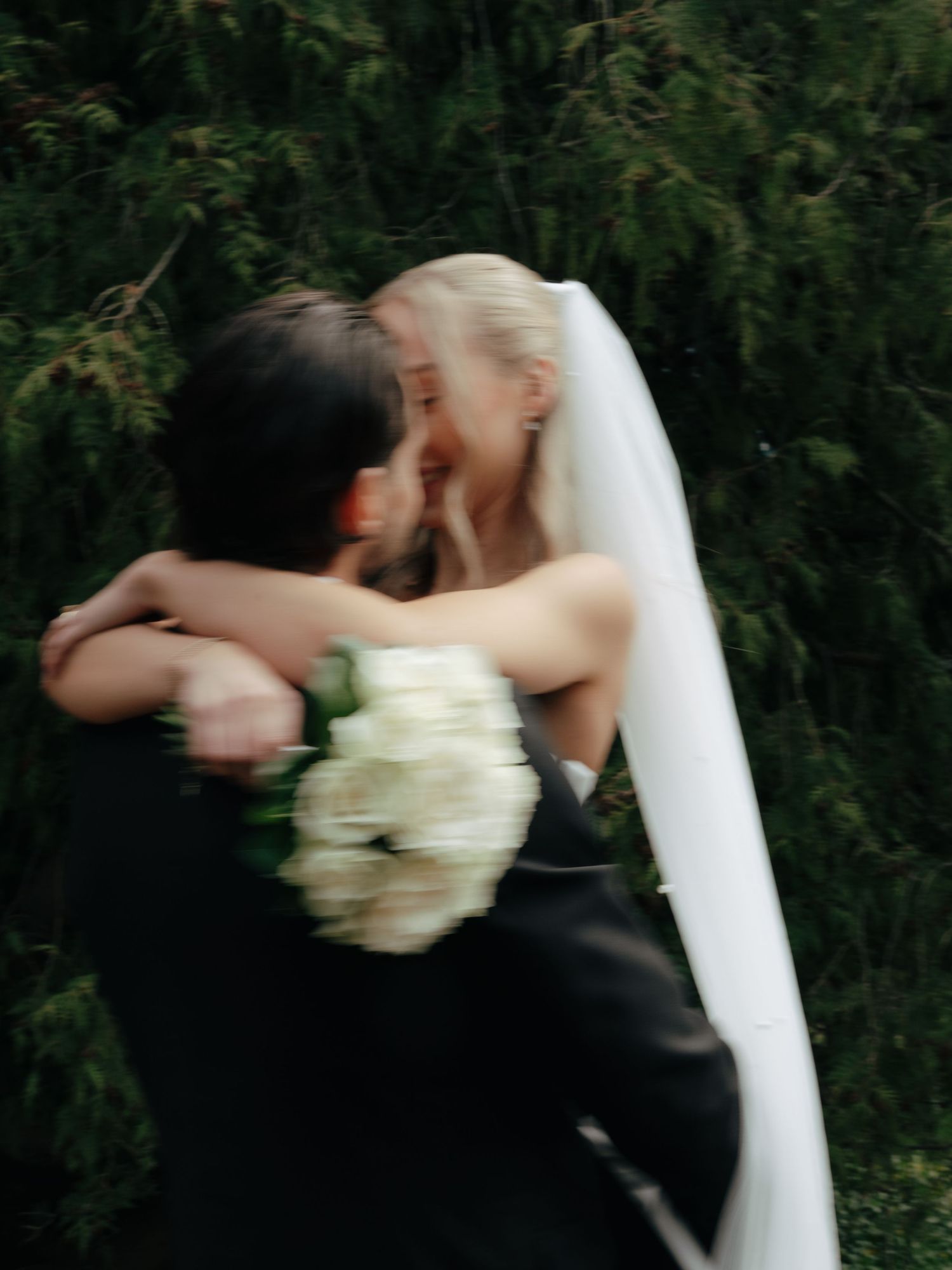 A bride in a white veil holding a bouquet embraces her groom in a black suit outdoors near lush green trees.