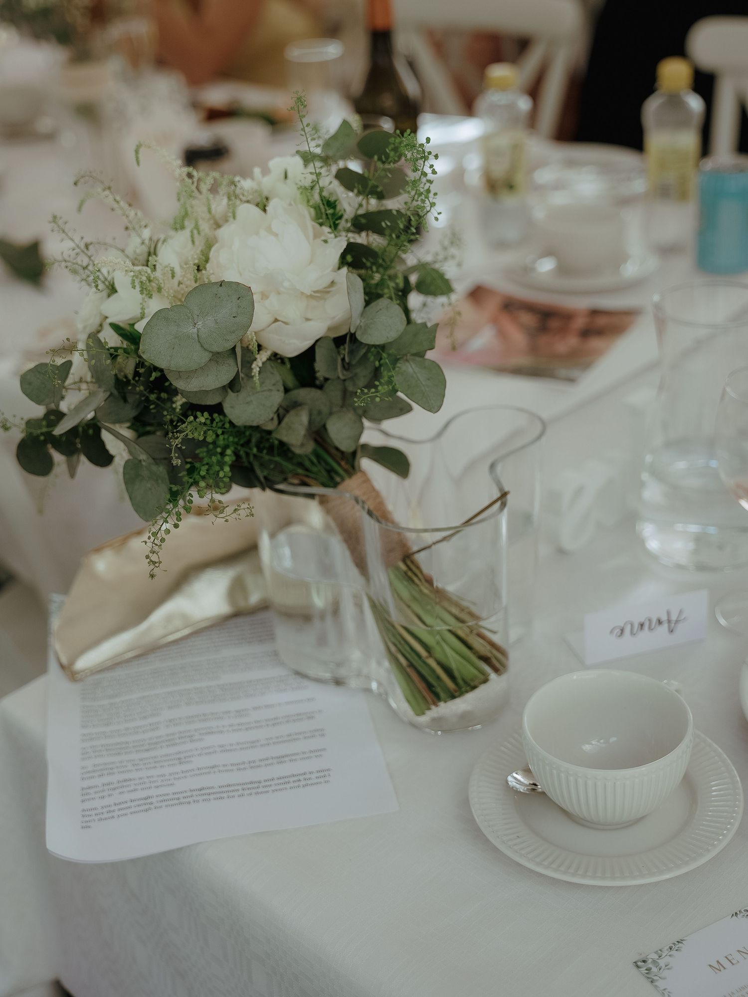 Elegant white floral wedding centerpiece with eucalyptus leaves sits on tables with white linens and place settings.