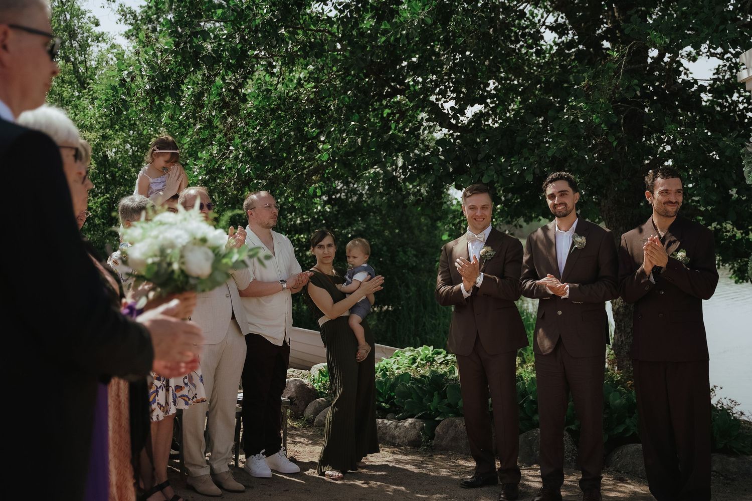 Wedding ceremony group stands in formation under trees with white floral arrangements.