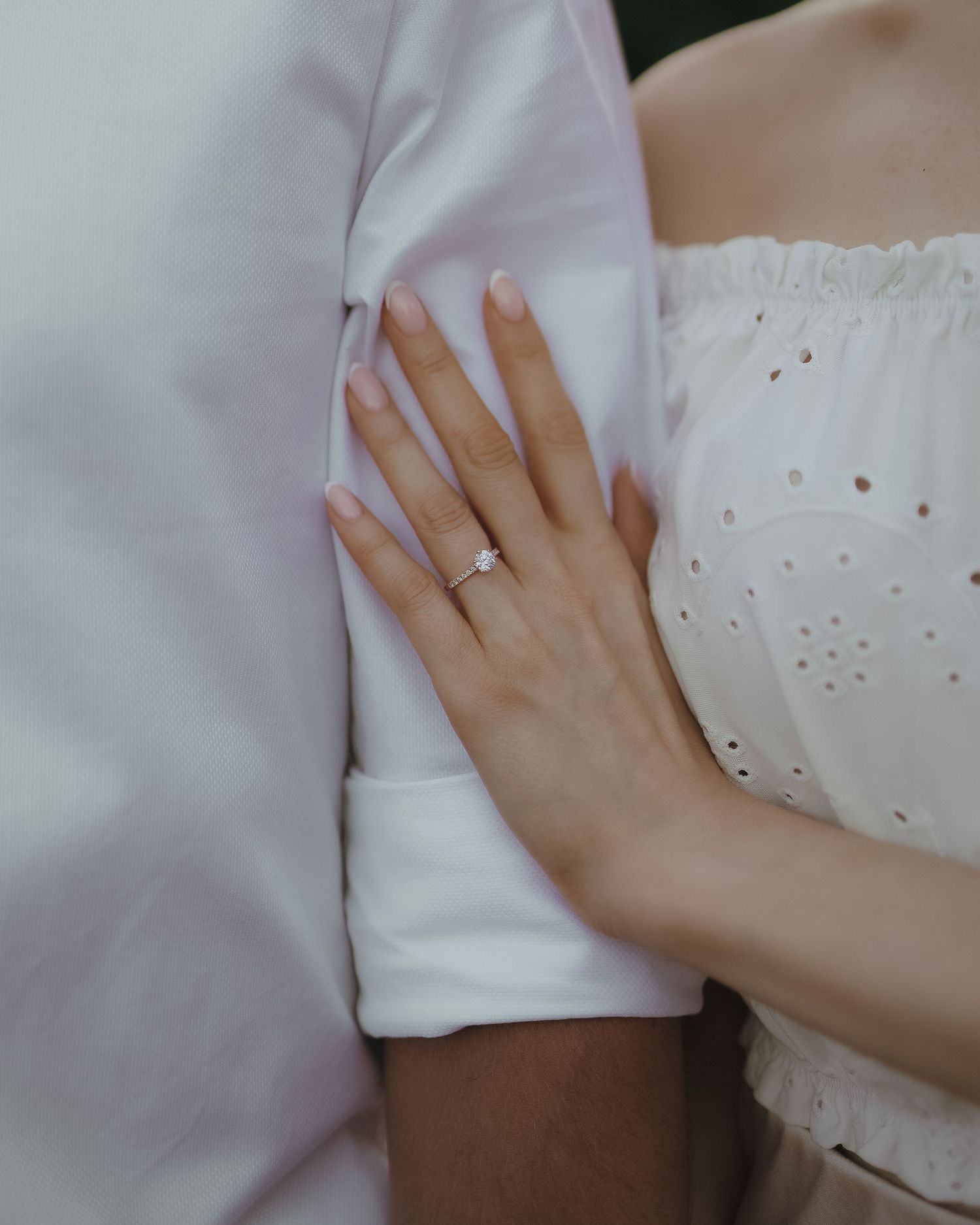 A close-up view of a hand with an engagement ring resting against a white fabric, showing both formal and casual attire.
