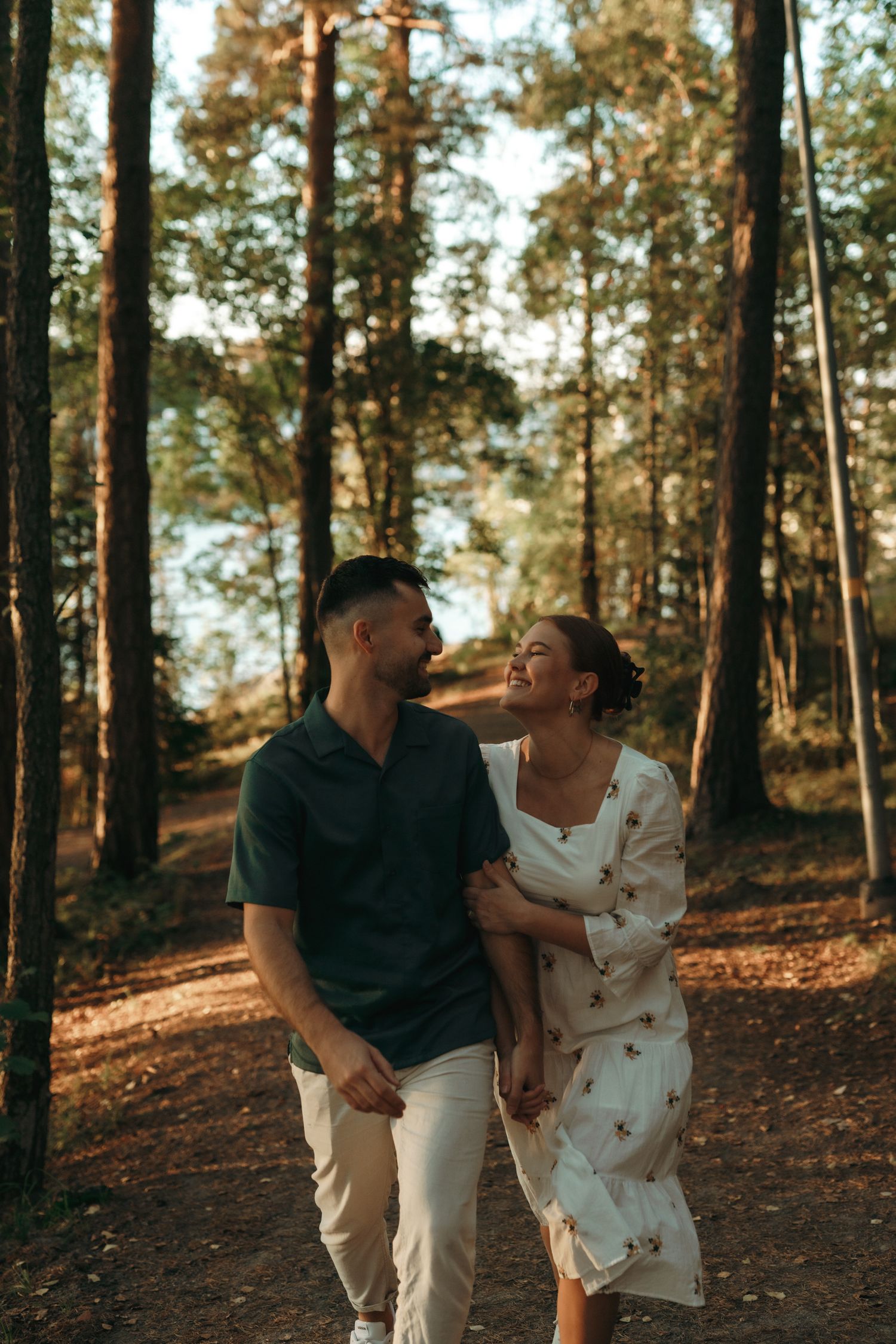 Happy couple walking together on a romantic forest path surrounded by tall pine trees at sunset.