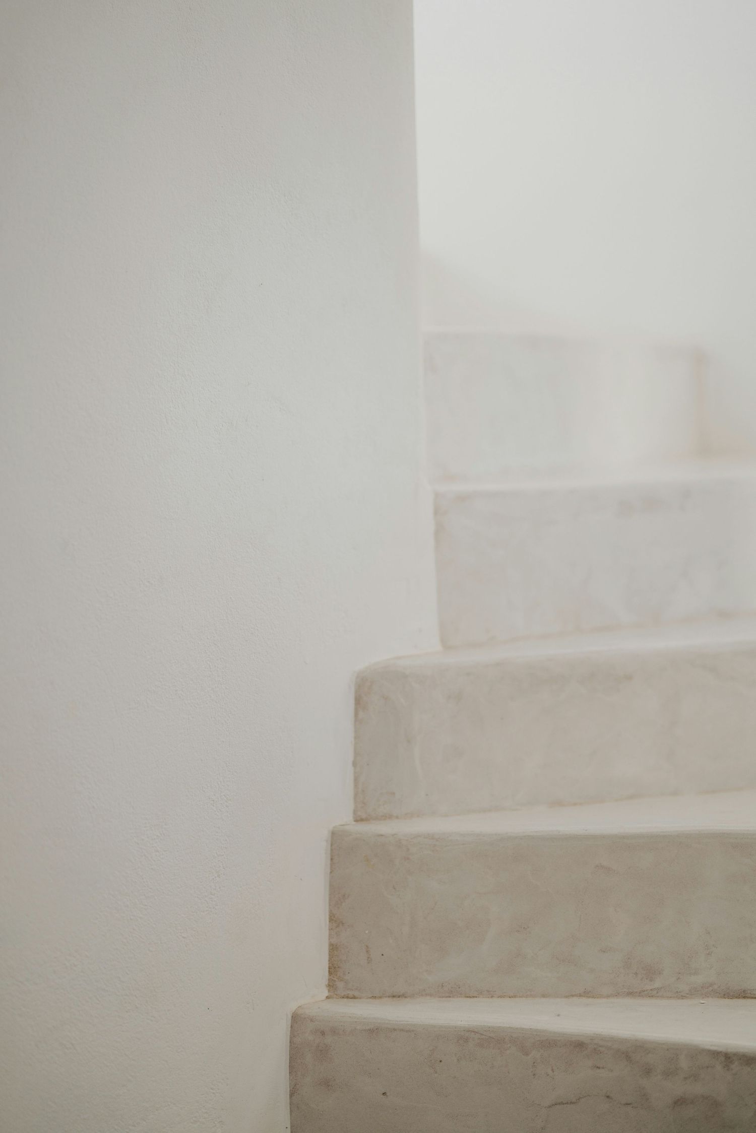 Minimalist white spiral staircase with concrete steps curving upward against a stark light-colored wall.