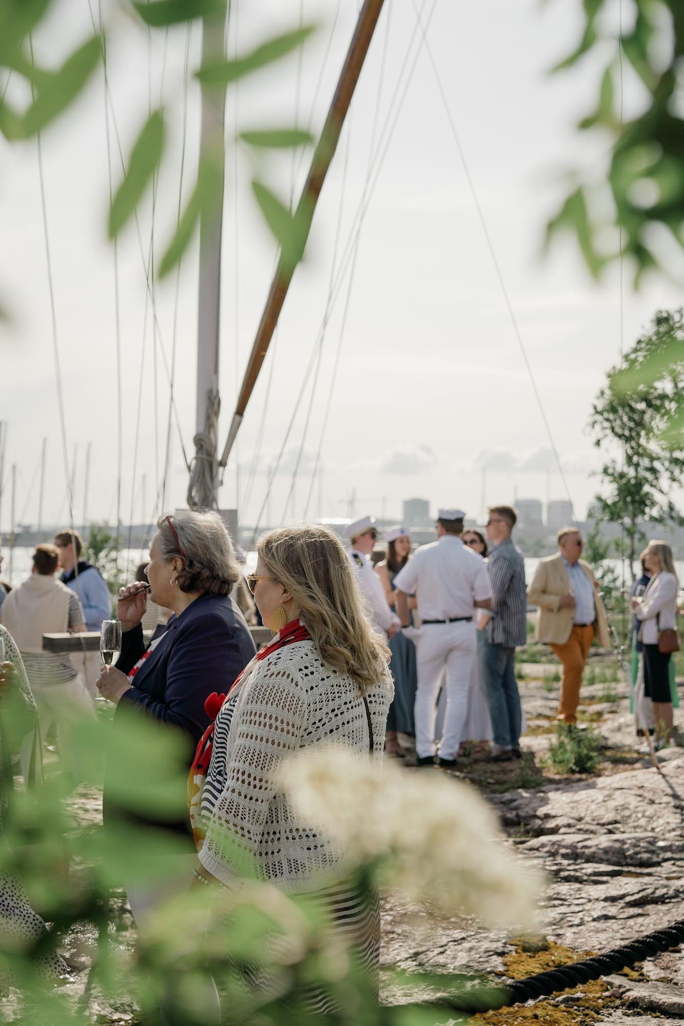 Guests gather near a sailboat mast by the waterfront on a sunny day, with lush greenery framing the scene.