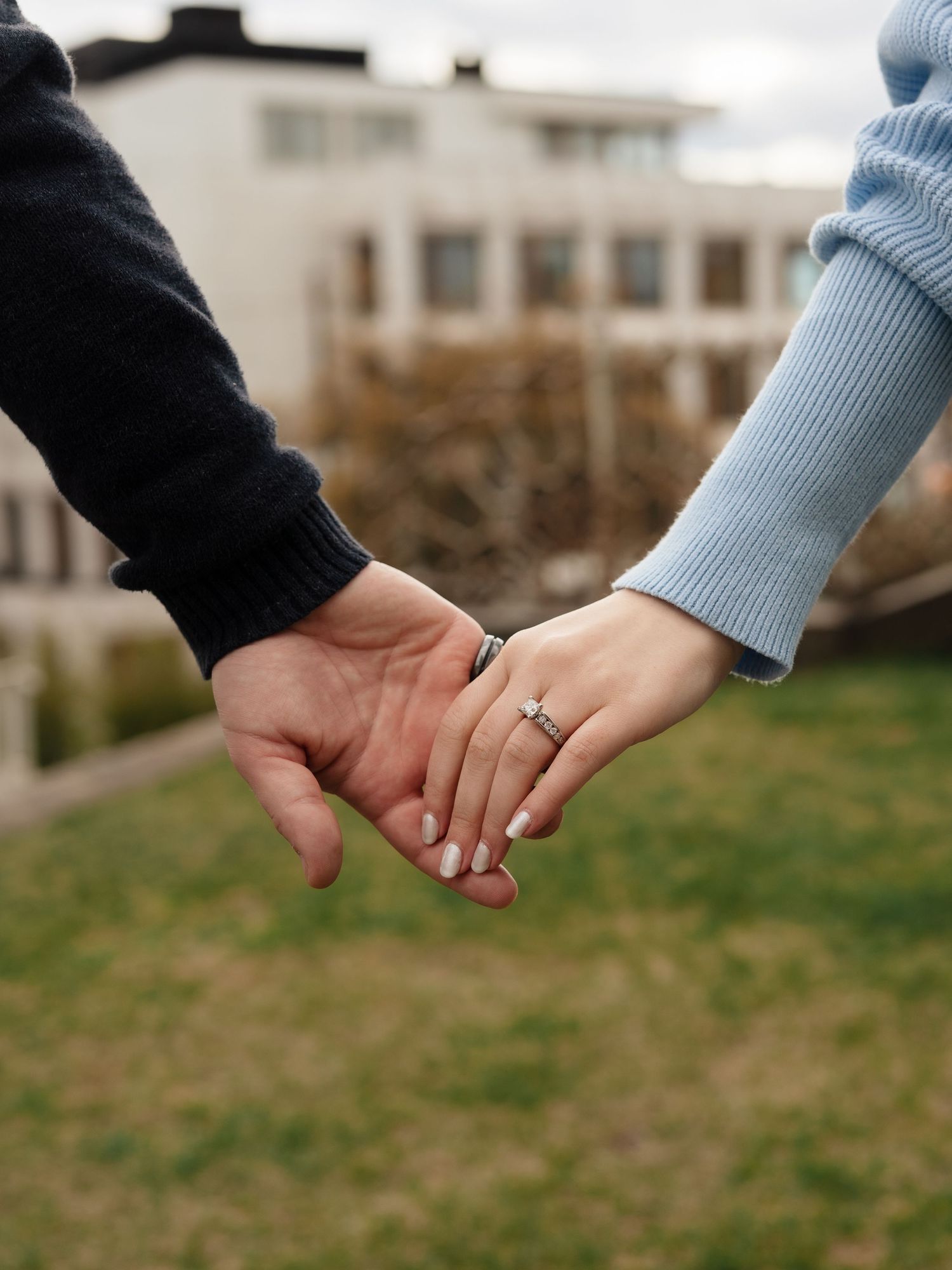 Couple holding hands outdoors, woman wearing engagement ring, blue sweater, romantic moment with blurred building background.