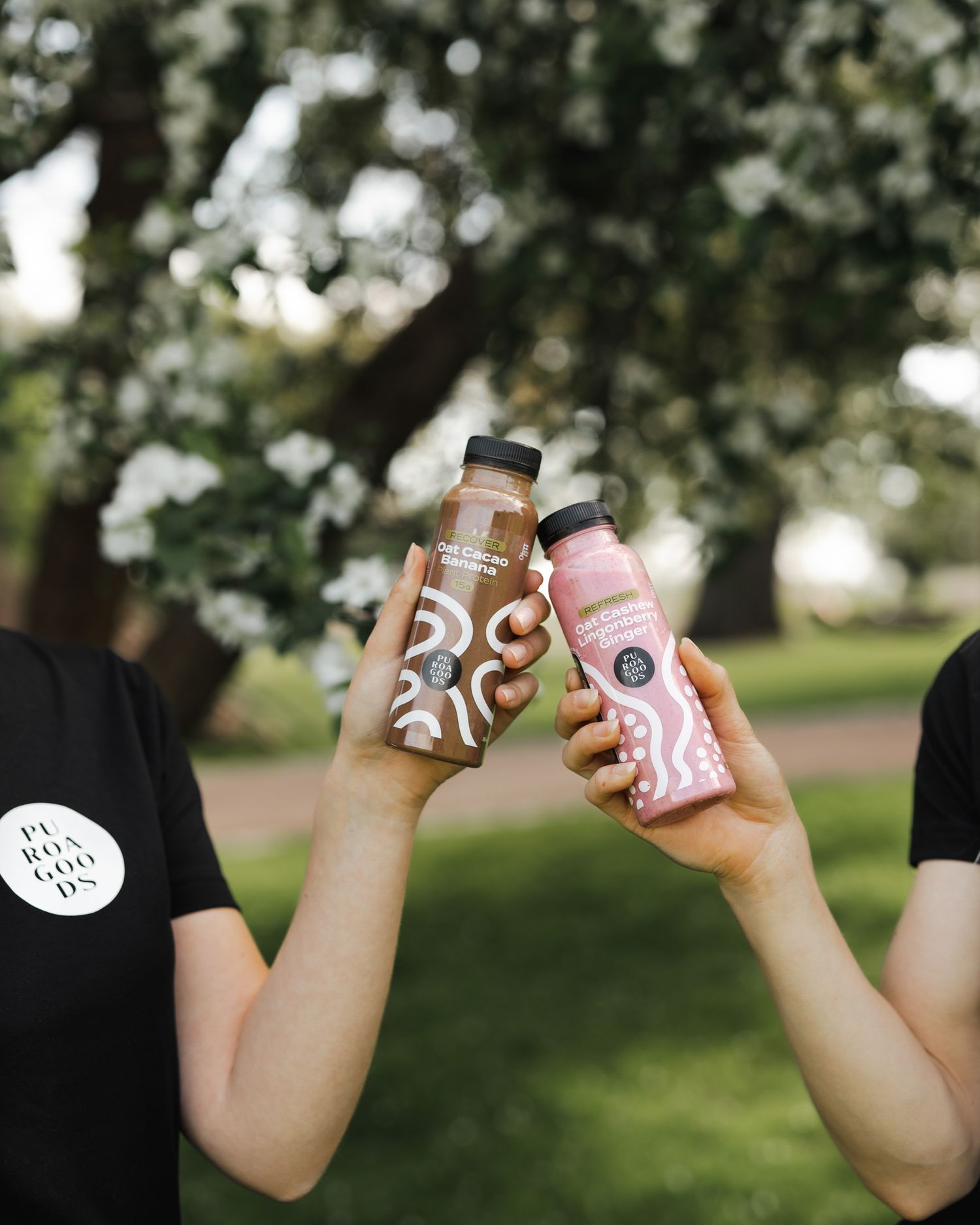 Two hands holding up glass bottles of refreshing pink and brown beverages in a garden setting with bokeh background.