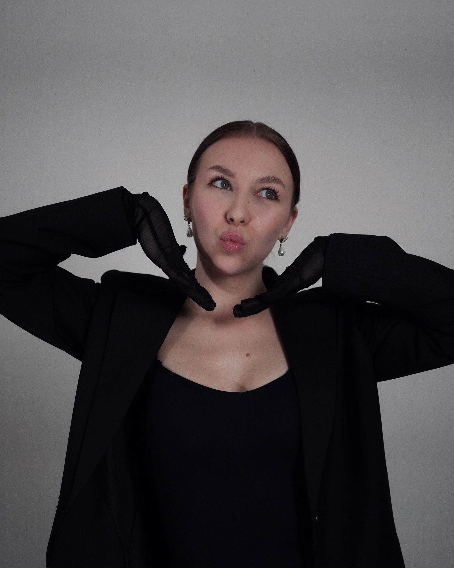 Someone in a black blazer and top makes a playful expression while posing against a gray background.