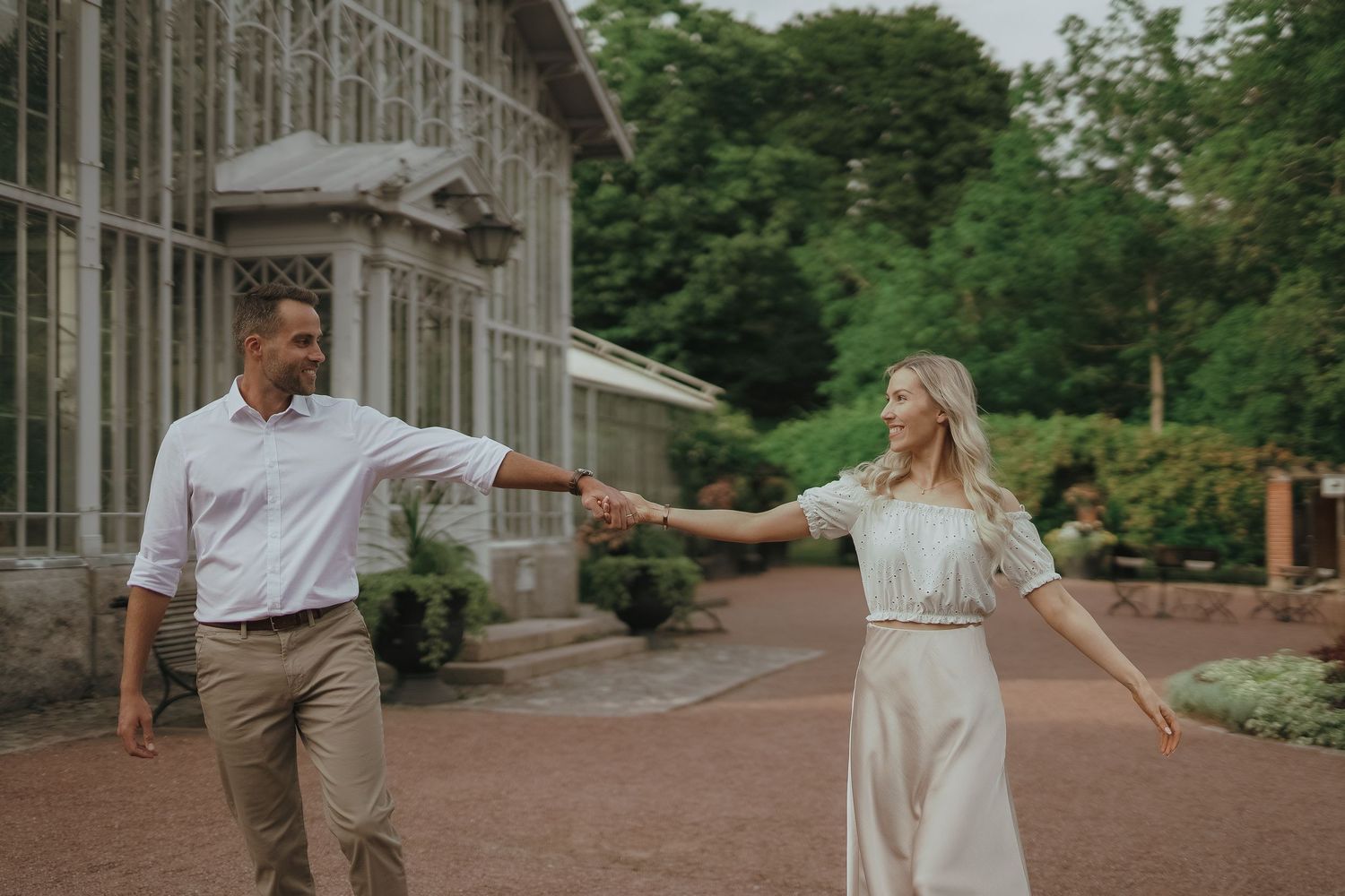 A couple in romantic evening attire dances together on a stone path surrounded by lush gardens and architecture.
