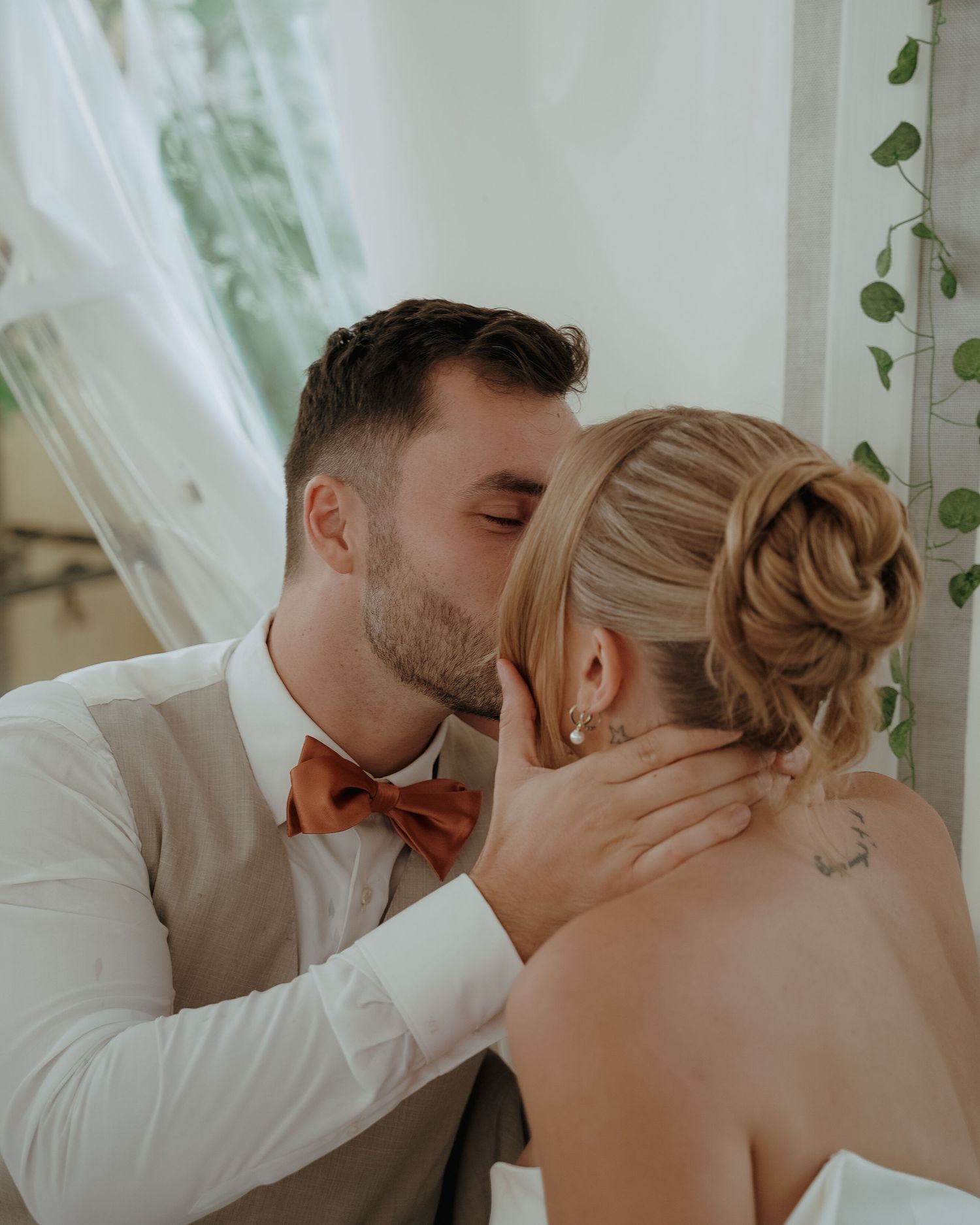 An intimate wedding moment captured with soft lighting and flowing white curtains and greenery in the background.