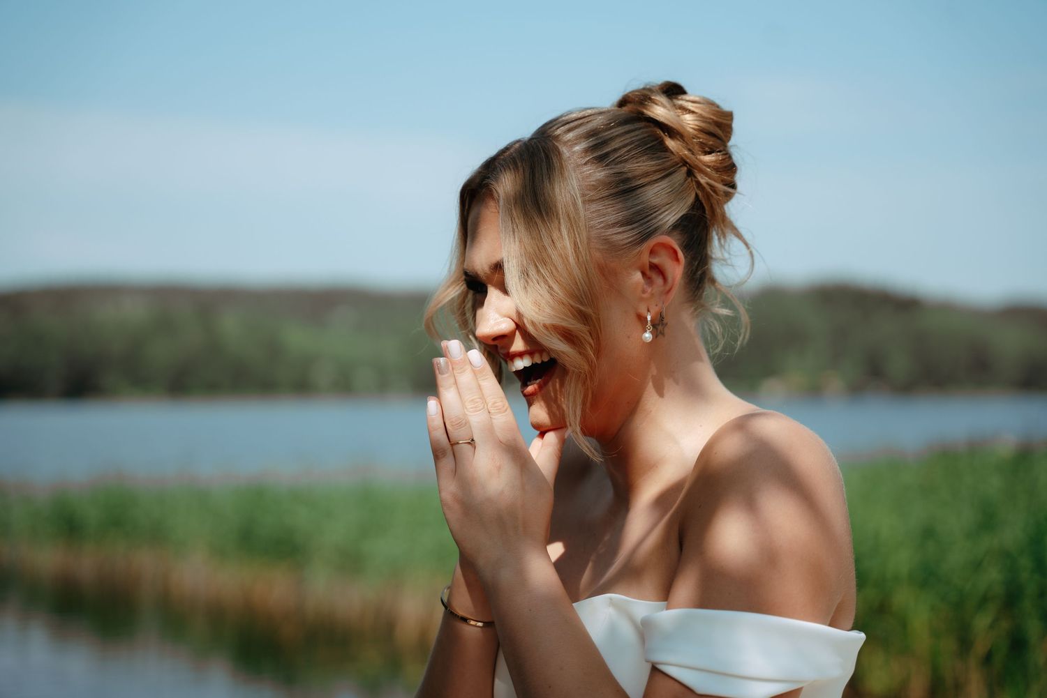 Someone in a white off-shoulder dress laughs candidly by a scenic lake and grassy shore on a sunny day.
